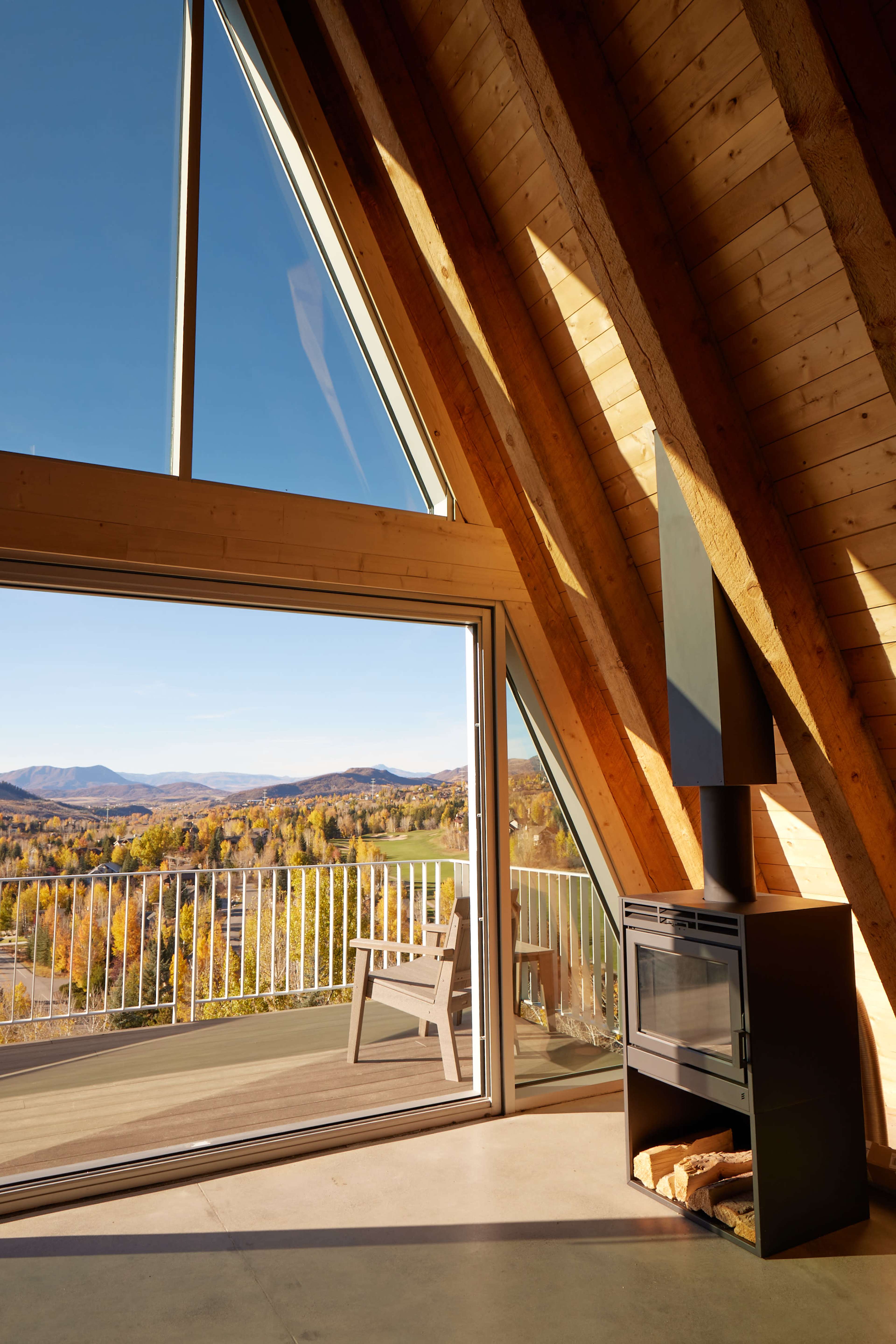 A modern triangular cabin interior features a wood stove and large windows offering a panoramic view of autumn landscapes outside.