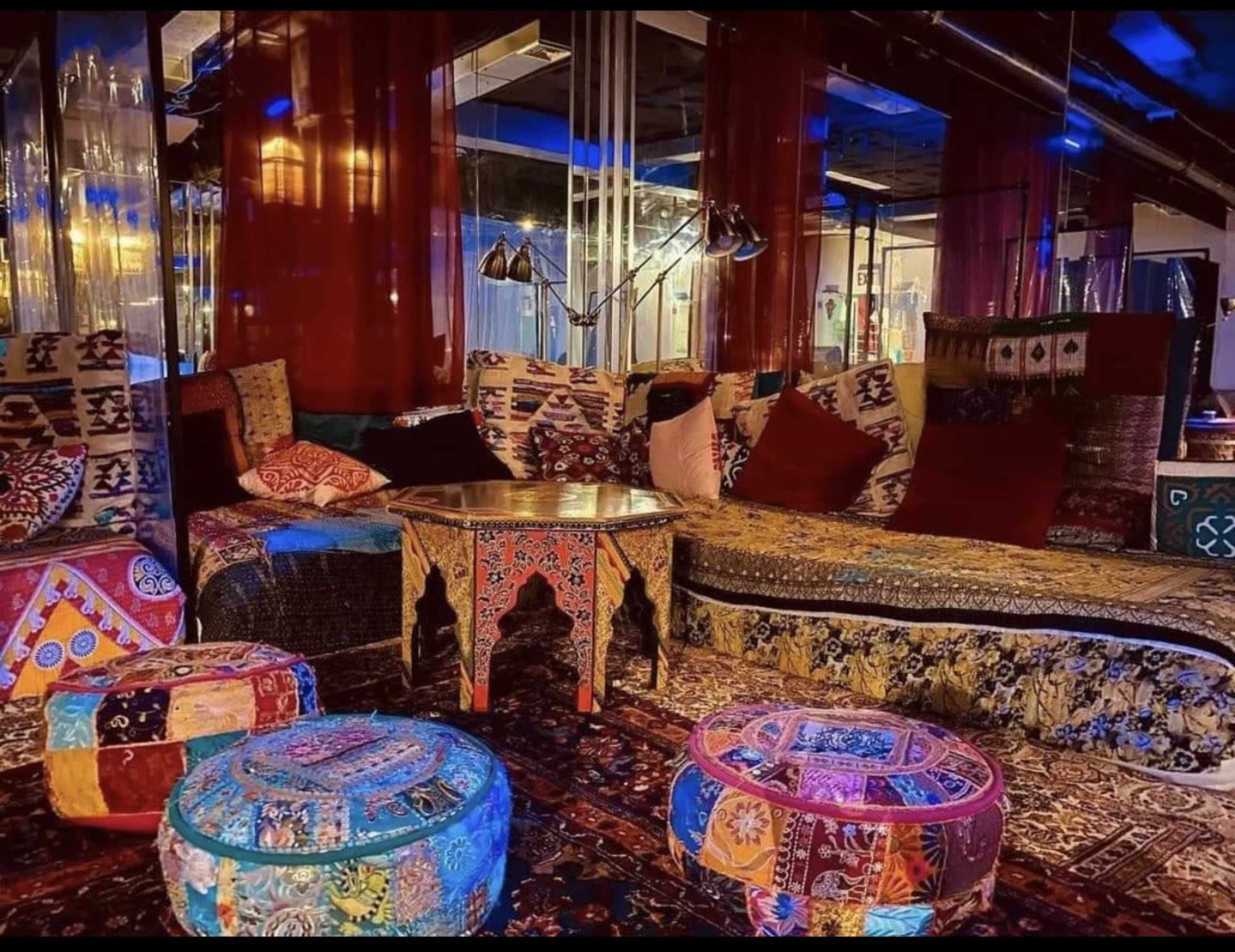 A cozy lounge area features intricately patterned textiles, colorful poufs, and a decorative table surrounded by plush seating.