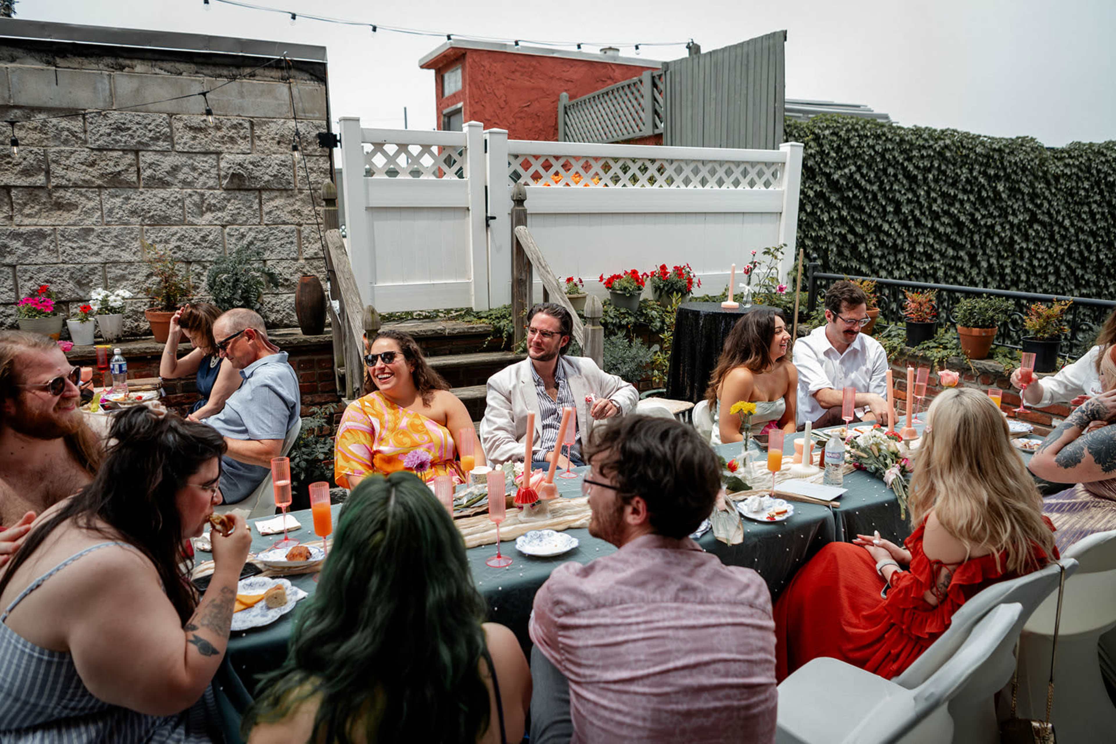 A group of people sits at a long table outdoors, enjoying a meal with drinks, surrounded by greenery and decorated with flowers.