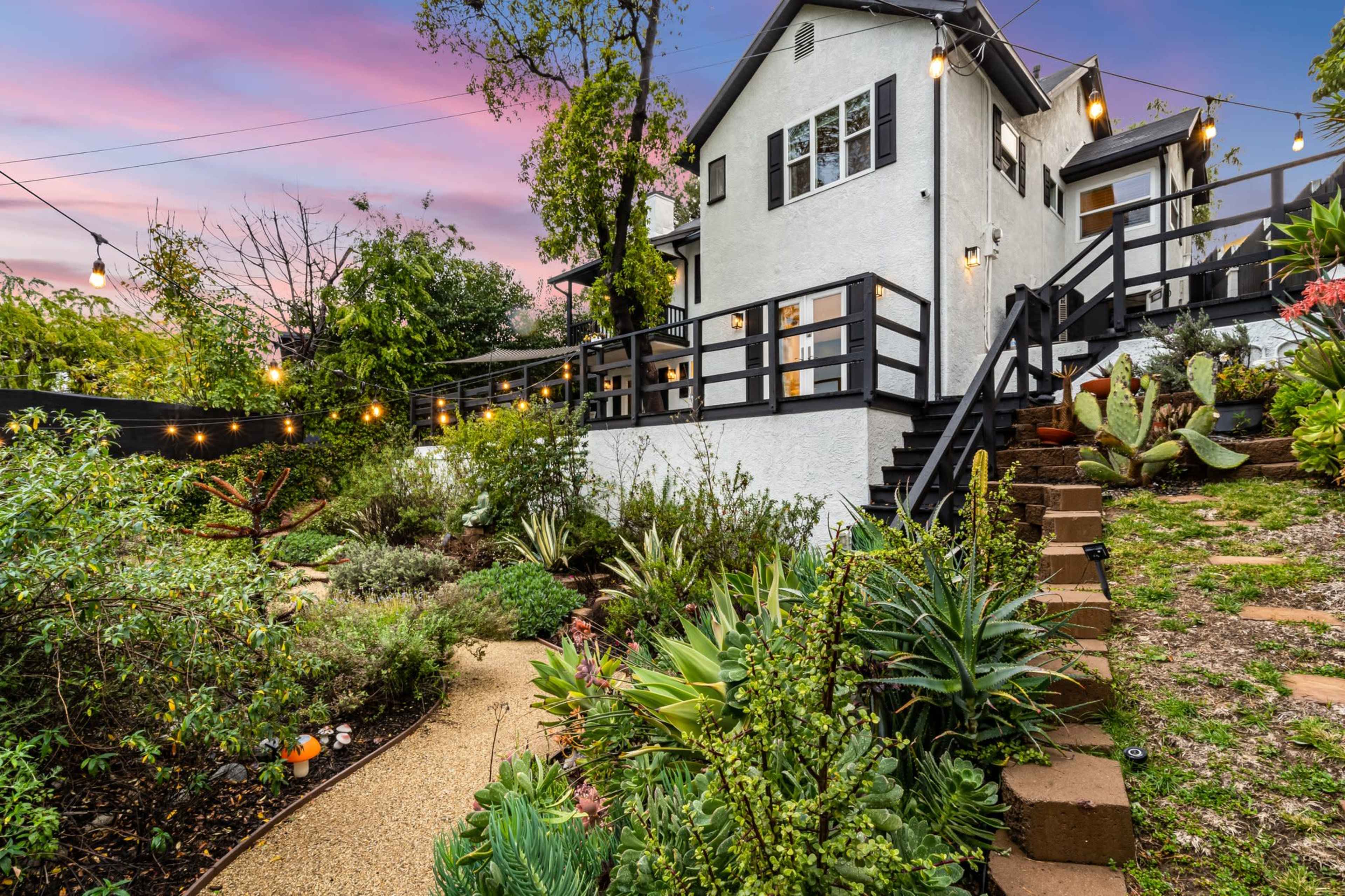 A house with a white exterior, surrounded by lush greenery and a pathway leading to a staircase, illuminated by string lights at dusk.