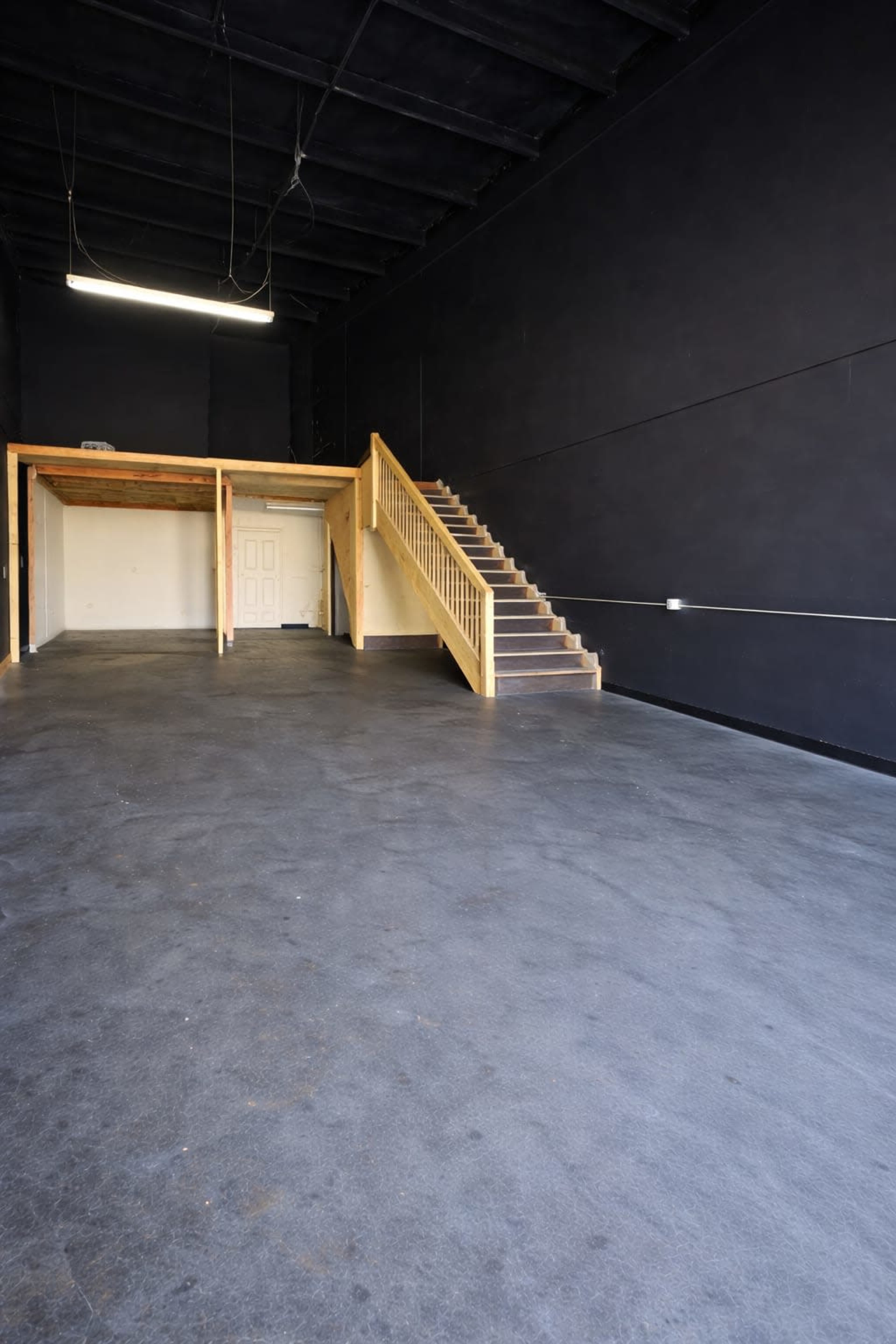 An empty room with a black wall, concrete floor, a staircase on one side, and a small elevated area at the back.