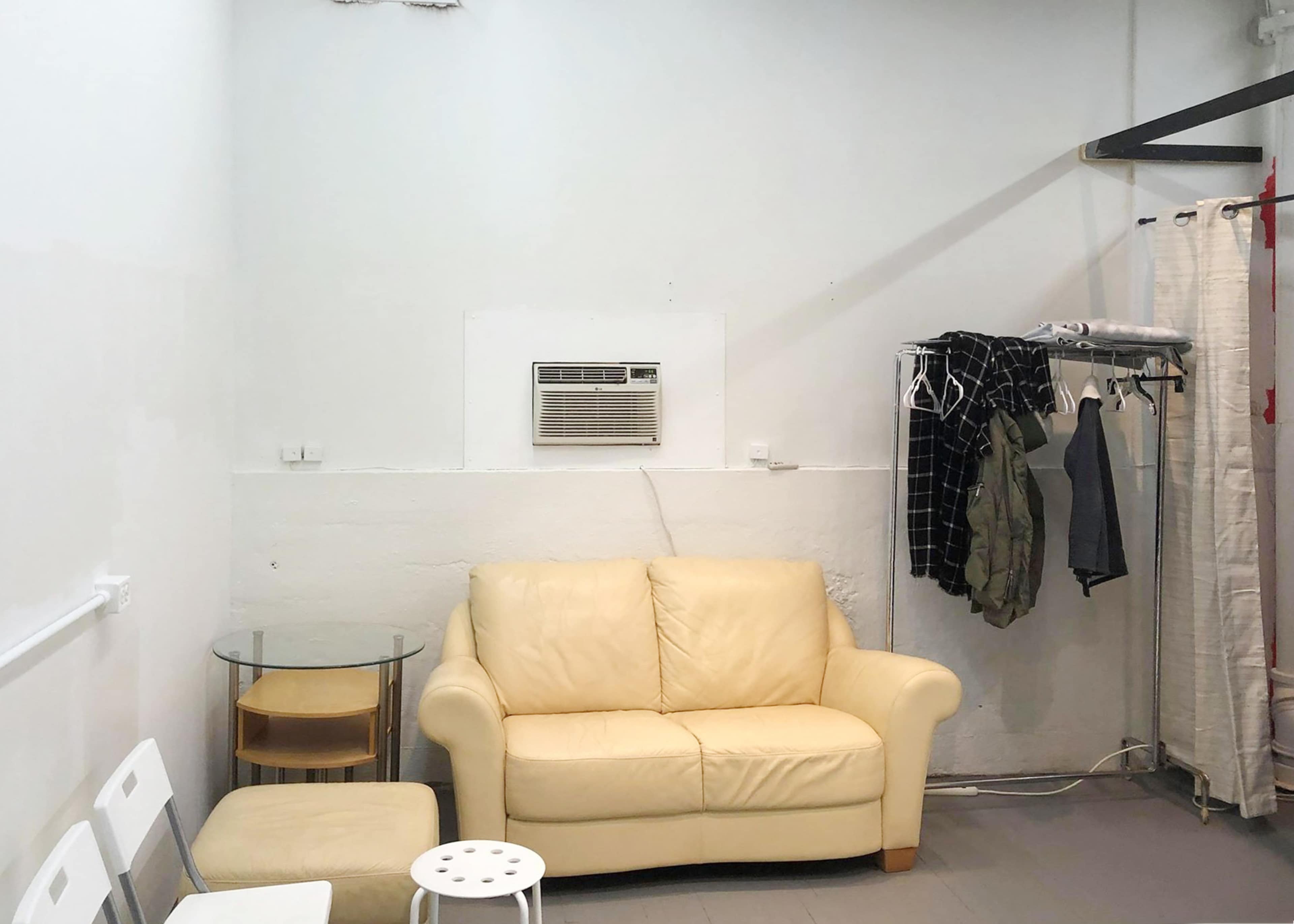 The image shows a small room featuring a light-colored couch, a glass side table, an air conditioning unit mounted on the wall, and a clothing rack with hanging garments.