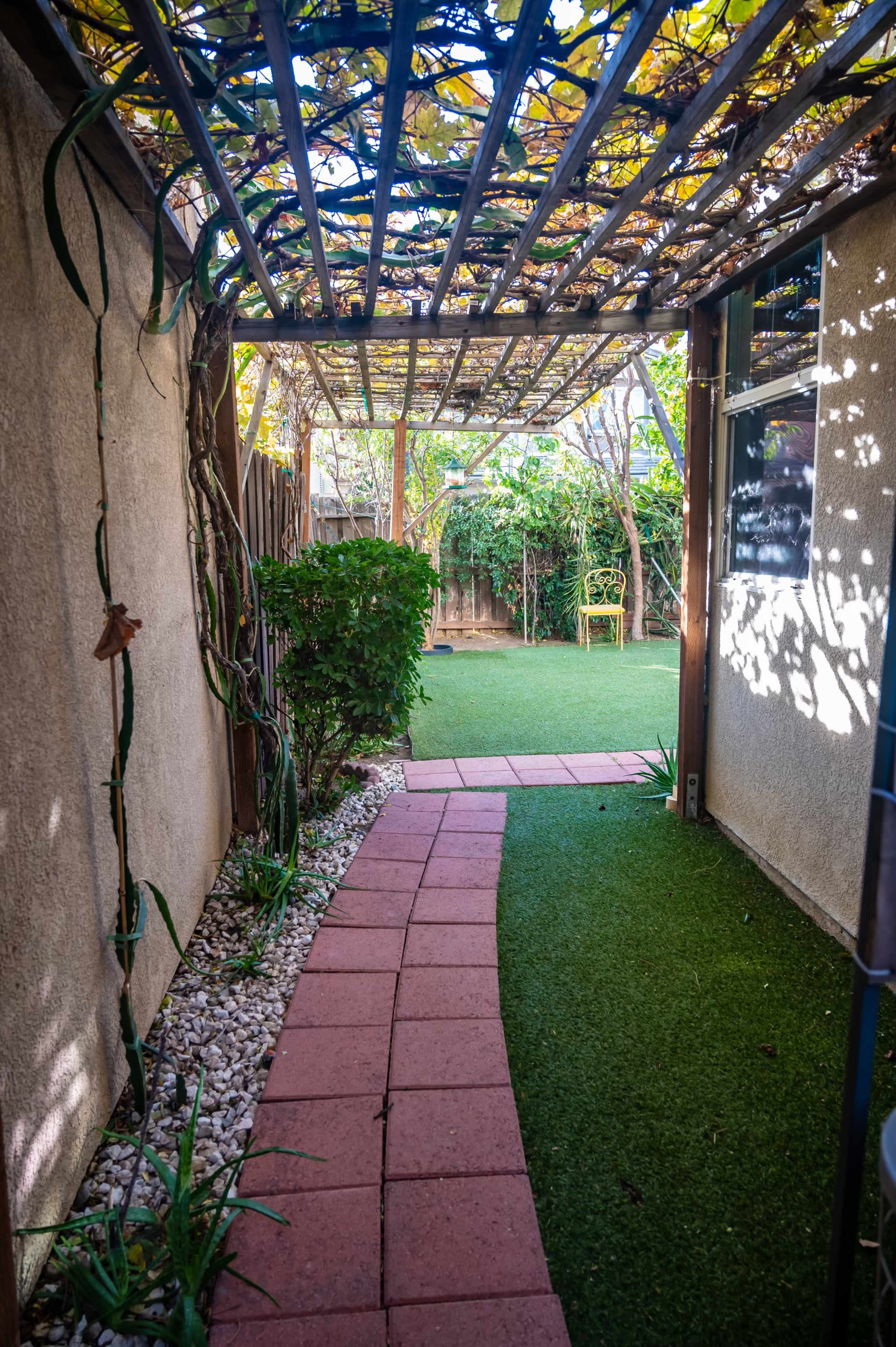A stone pathway leads through a trellis-covered walkway toward a green lawn surrounded by shrubs.