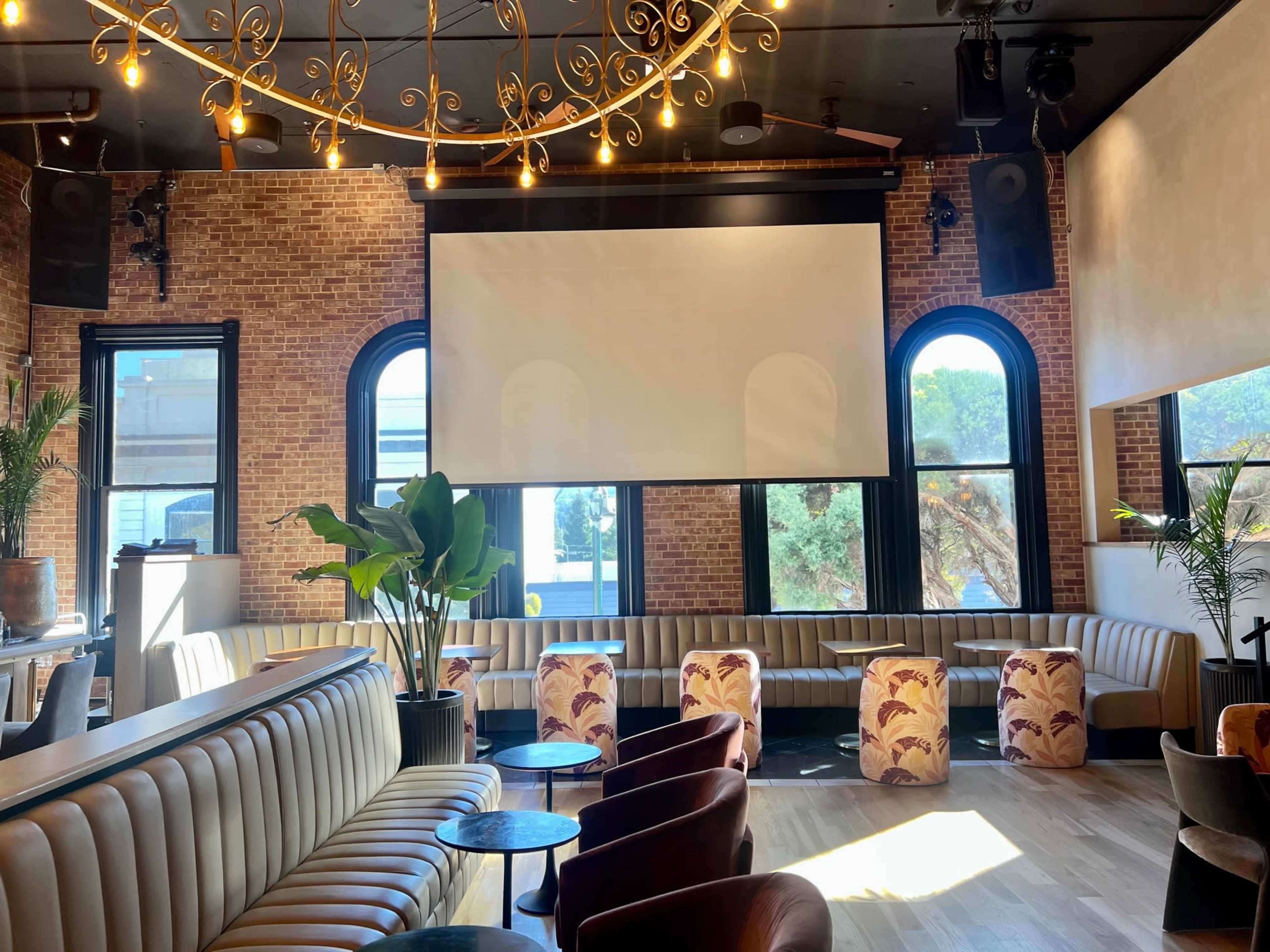 The image shows a modern lounge area featuring curved seating, a large projection screen, and decorative lighting in a brick-walled interior.