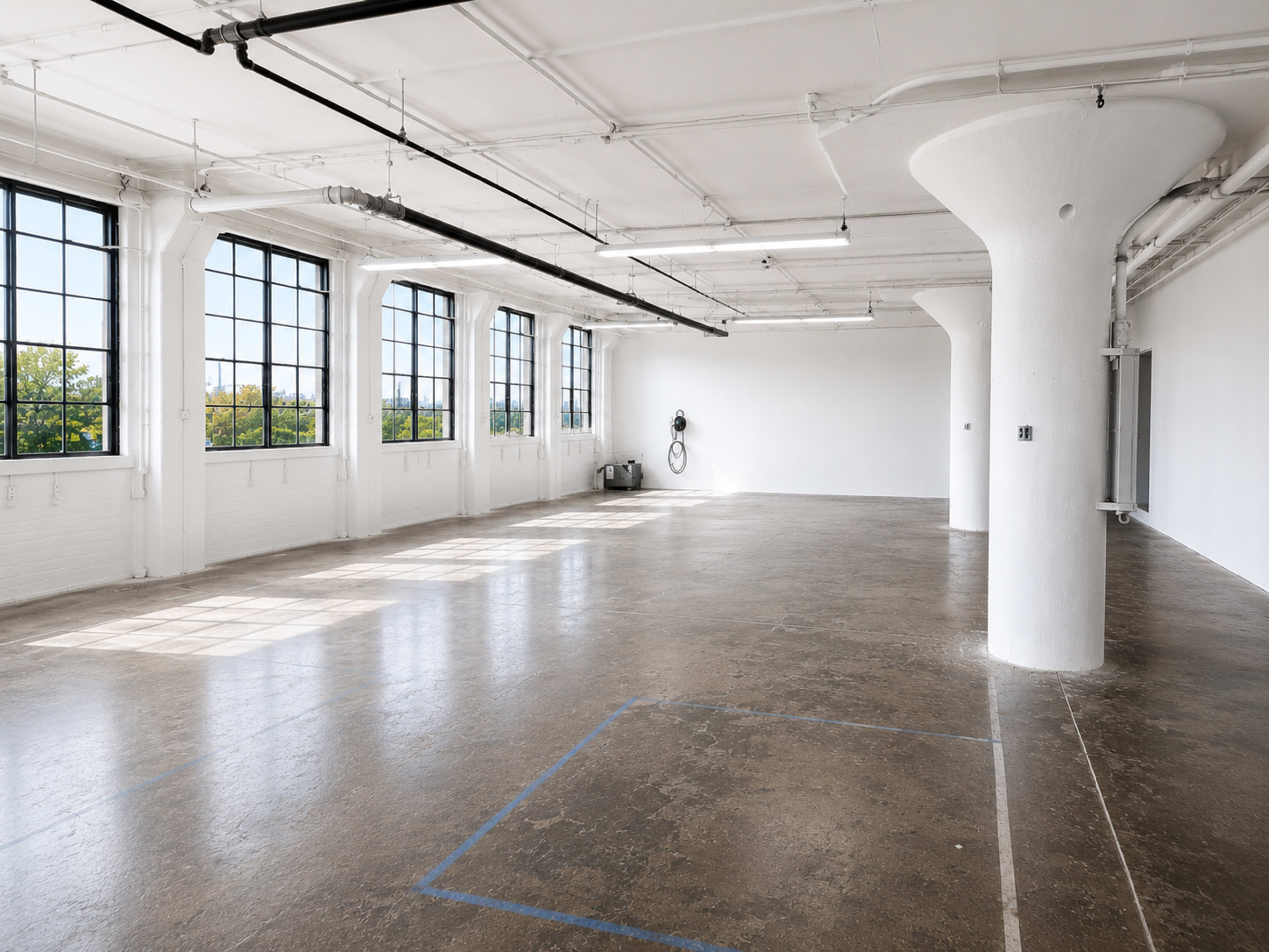 The image shows a spacious, industrial-style room with large windows, a concrete floor, and a central white column.