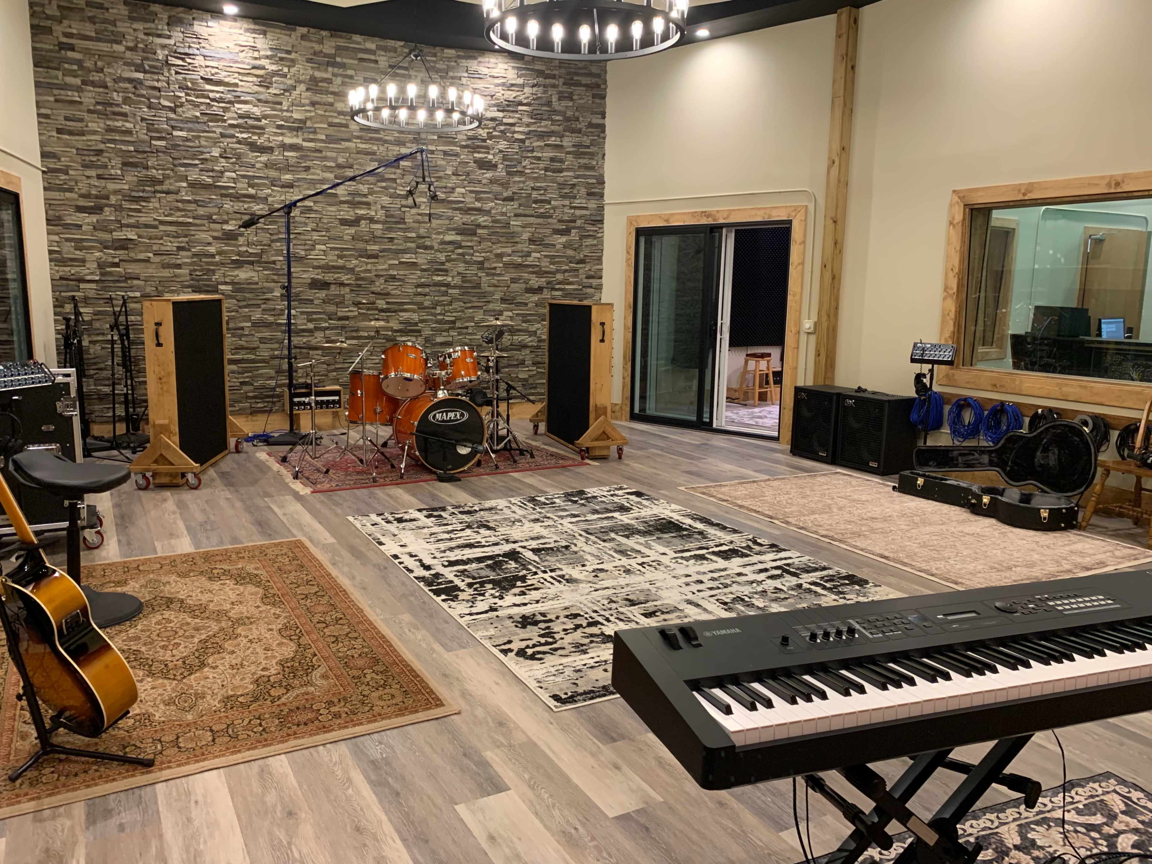 The image shows a music studio featuring a drum set, a keyboard, amplifiers, guitars, and spacious areas with rugs and a stone accent wall.
