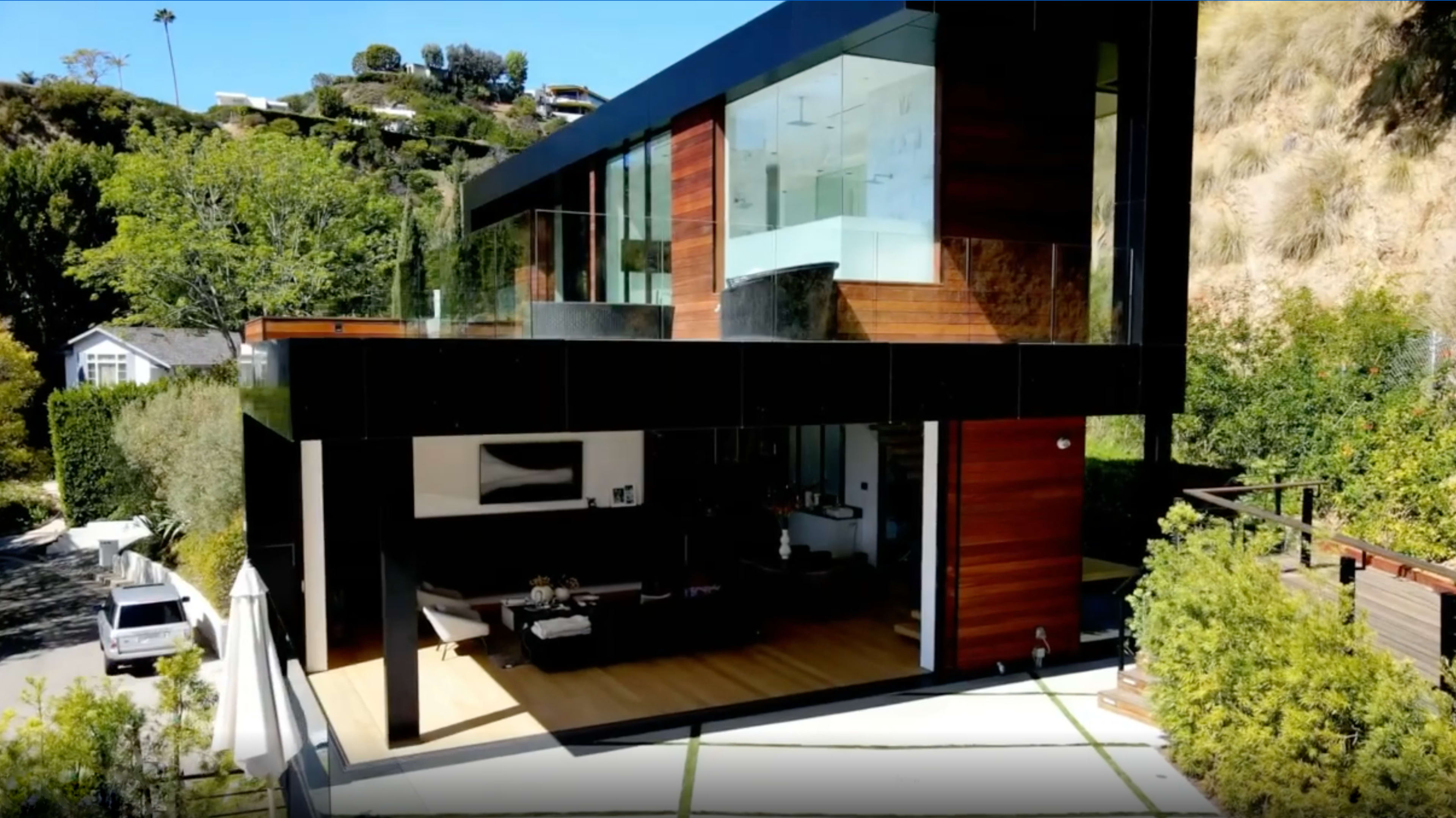 A modern two-story house with large glass windows and wooden paneling, surrounded by greenery.