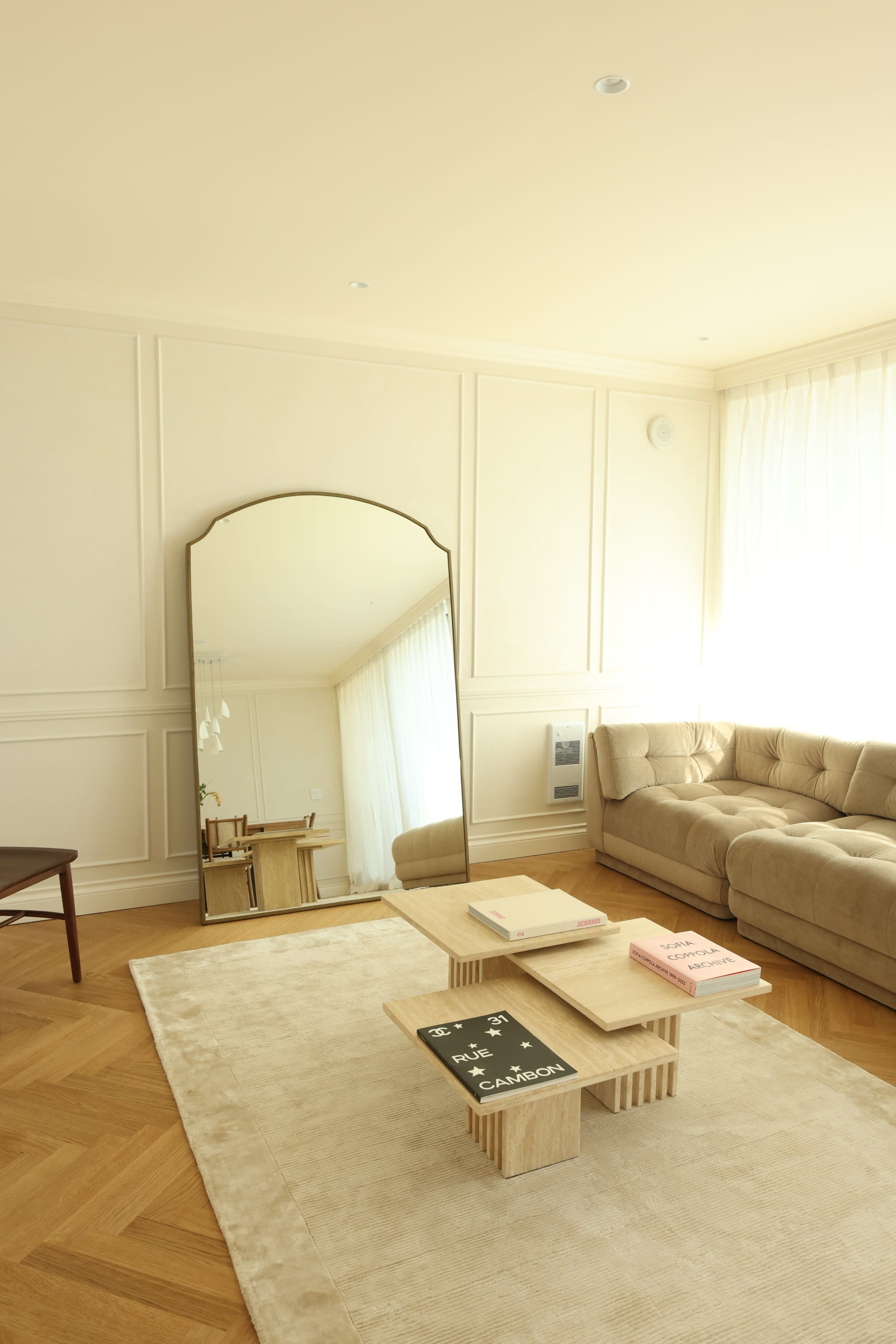 The image shows a modern living room with a beige sofa, a large mirror, and a wooden coffee table on a light-colored rug.