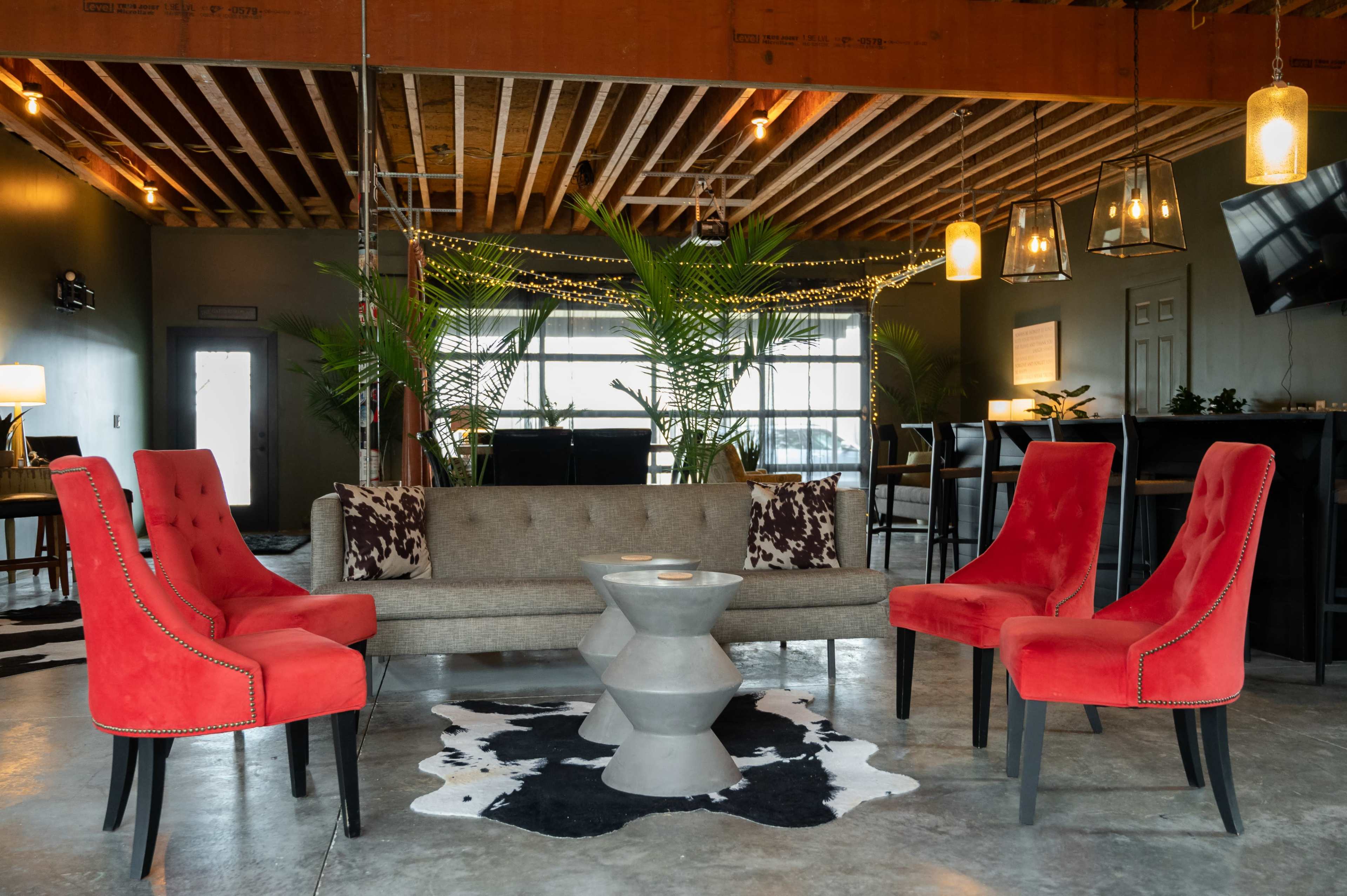 A modern lounge area featuring a gray sofa, four red chairs, and a geometric coffee table, all situated on a black and white cowhide rug beneath exposed wooden beams and ambient lighting.