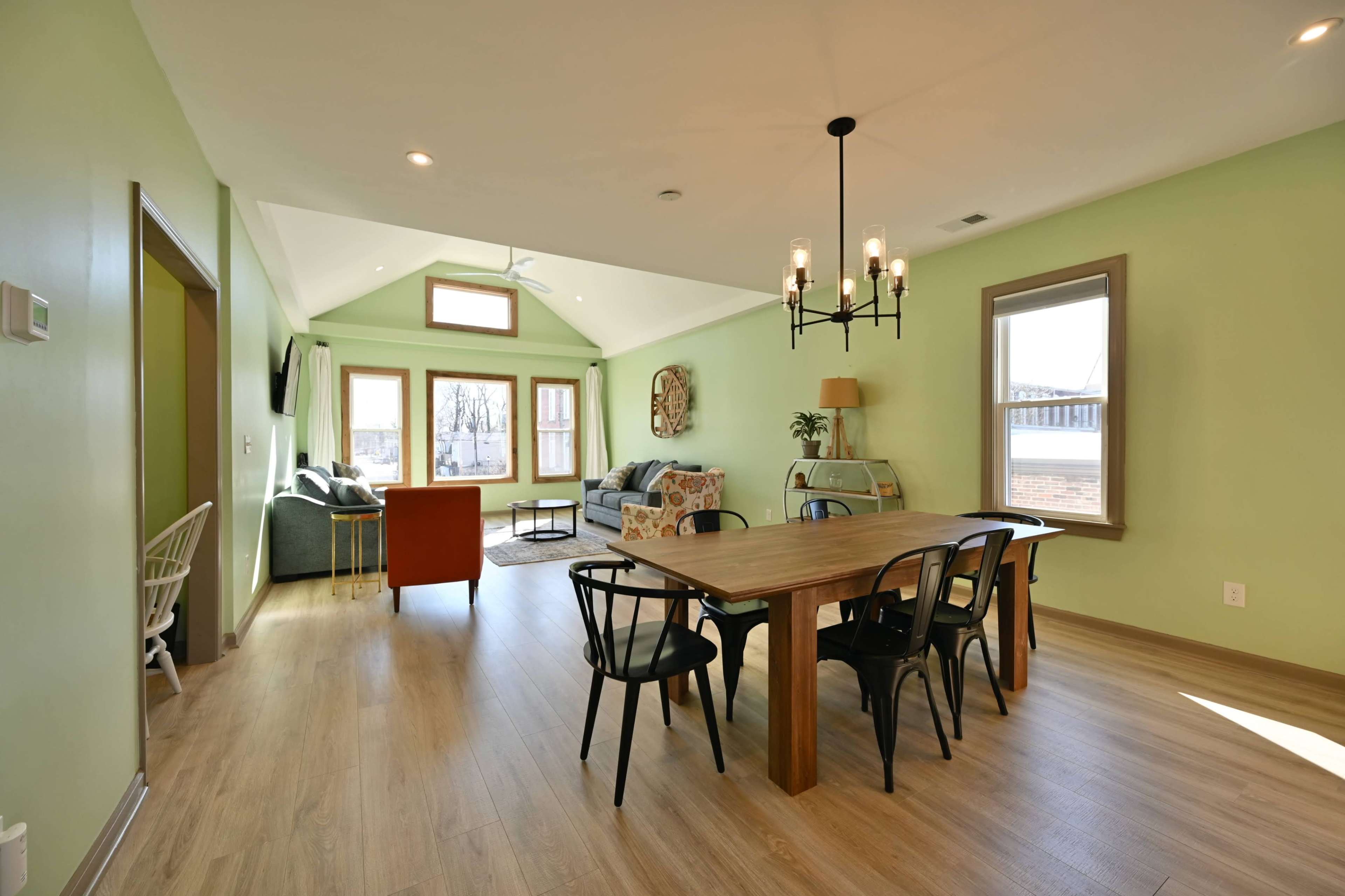 The interior of a modern room features a dining area with a wooden table and black chairs, a living space with a couch and armchair, and large windows allowing natural light.