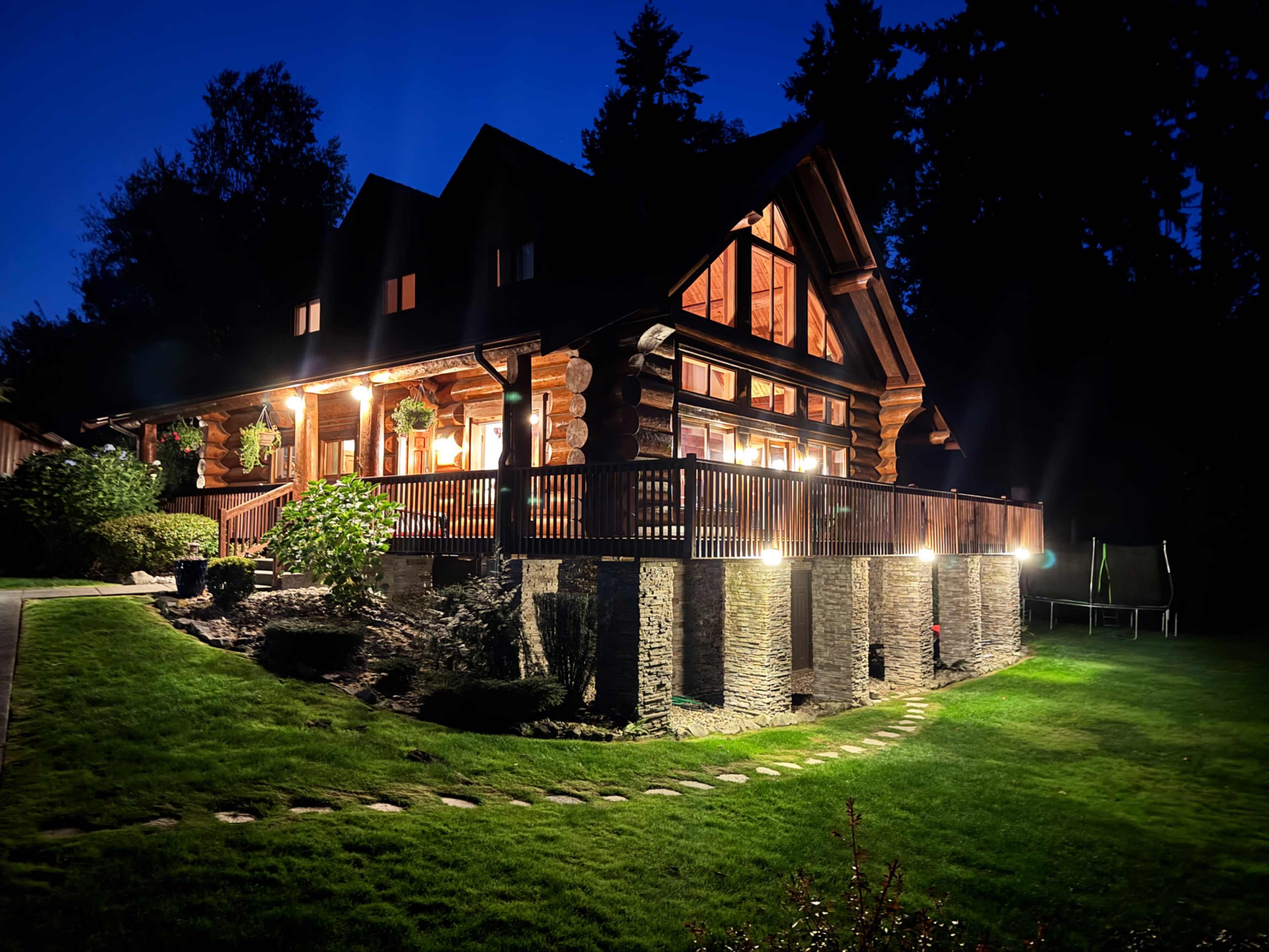 A log cabin with large windows is illuminated at night, surrounded by neatly manicured lawns and garden features.