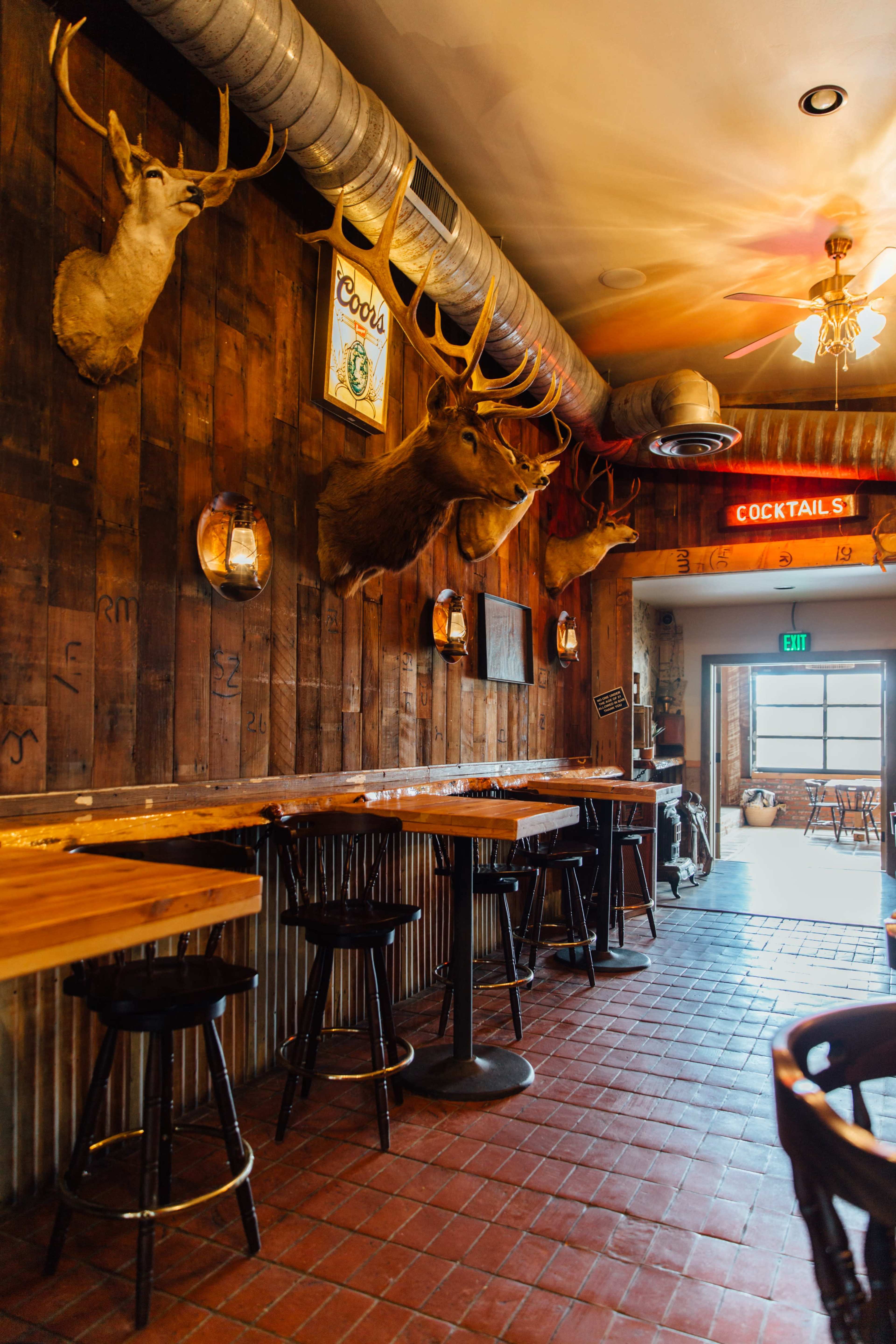 The interior of a rustic bar features wooden walls adorned with mounted deer heads and a row of high stools at a wooden counter.