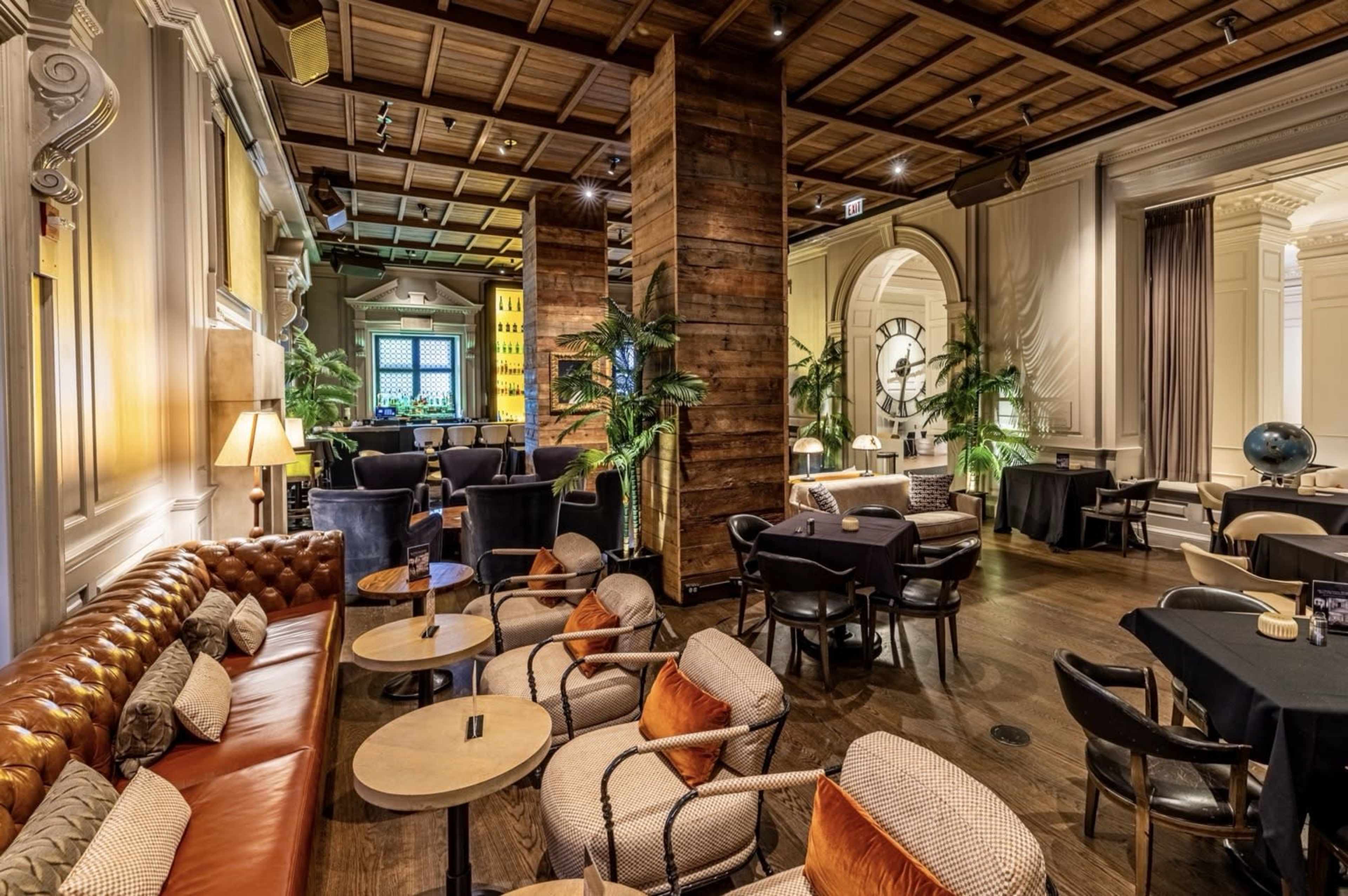 The image shows a stylish lounge area featuring a mix of brown leather seating, wooden columns, and potted plants, with tables arranged for patrons.