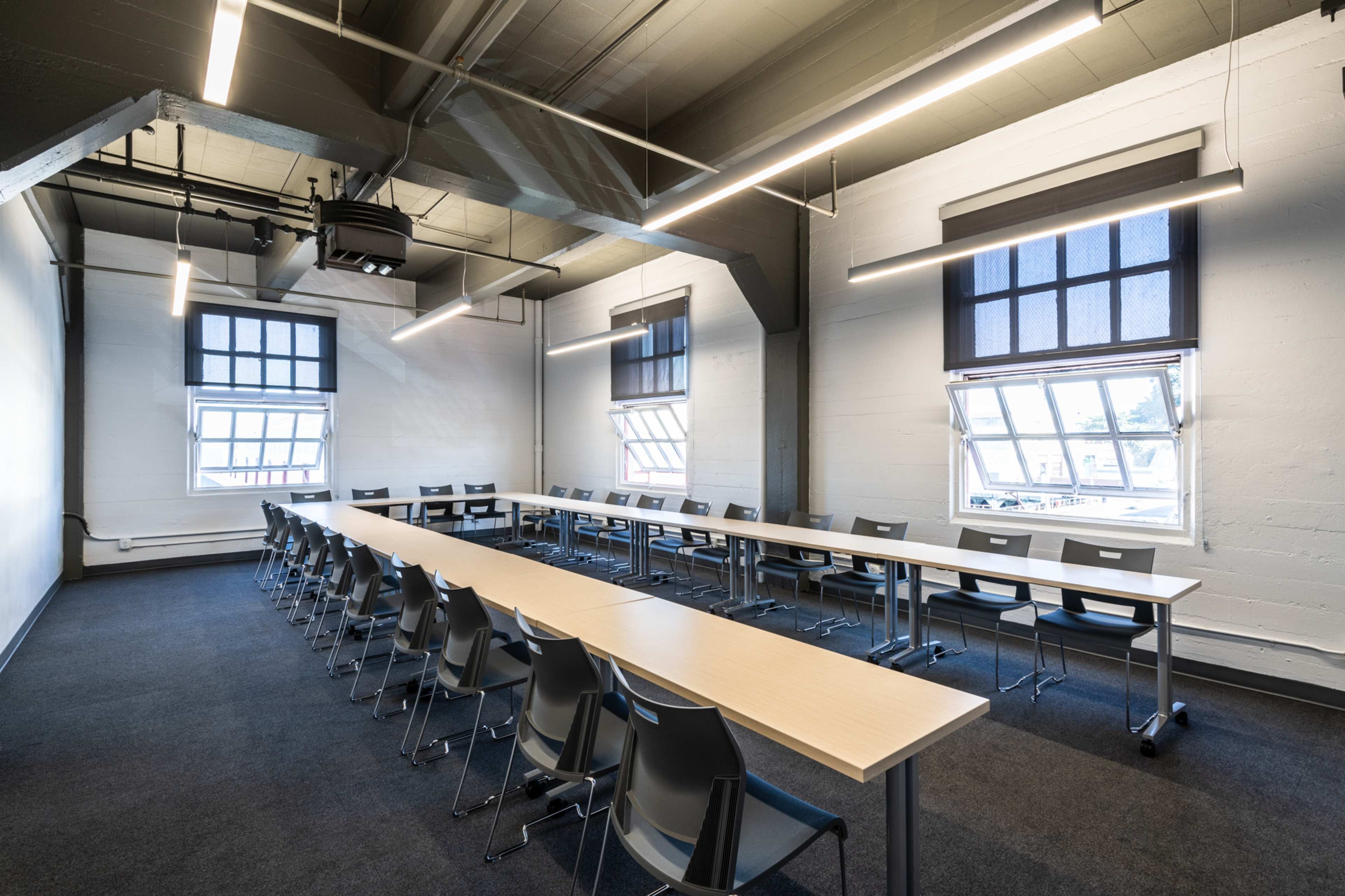 A spacious classroom features a long table with multiple chairs arranged around it and large windows allowing natural light.