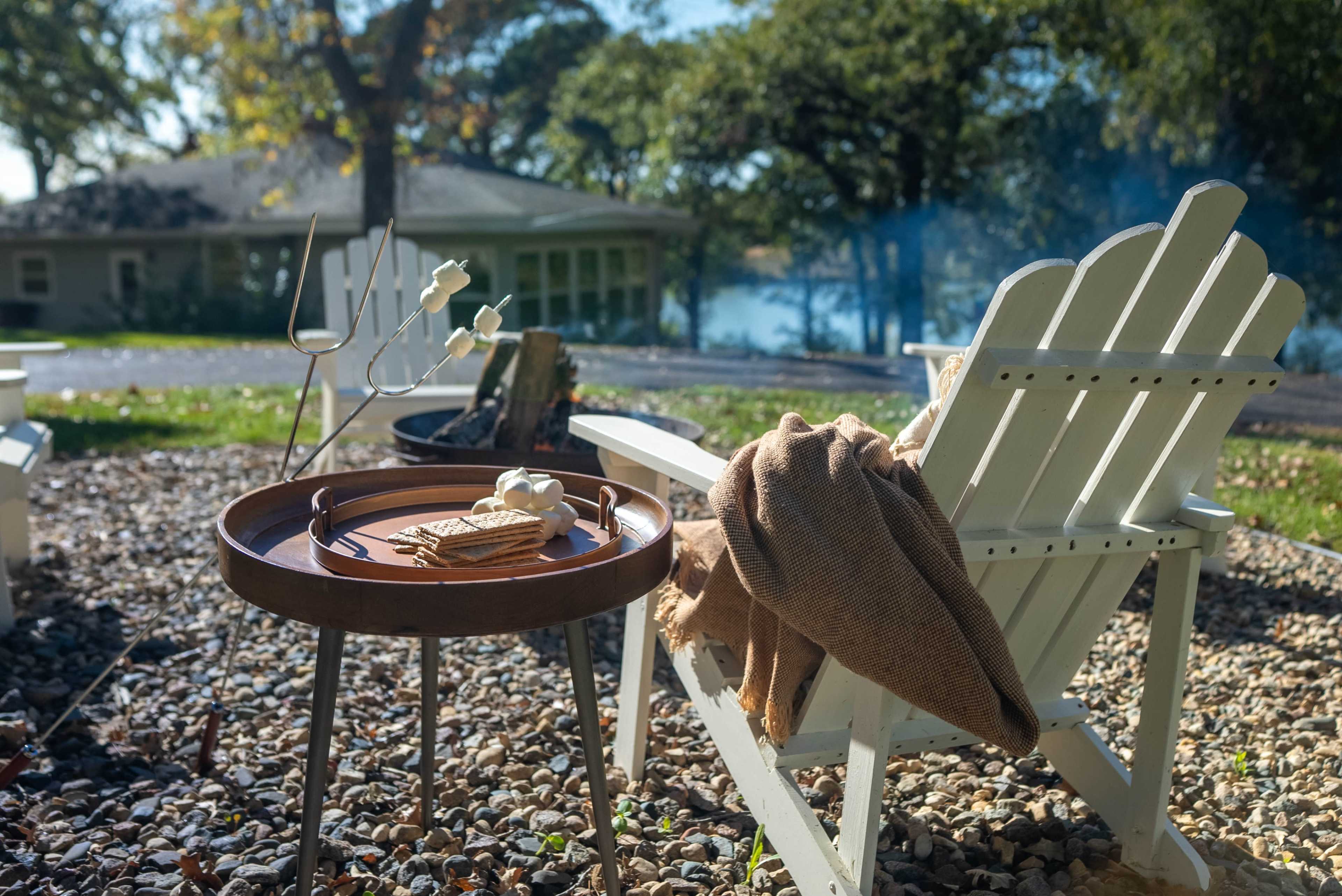 A wooden tray with snacks sits on a small table next to a white Adirondack chair, set in a gravel area near a lake.