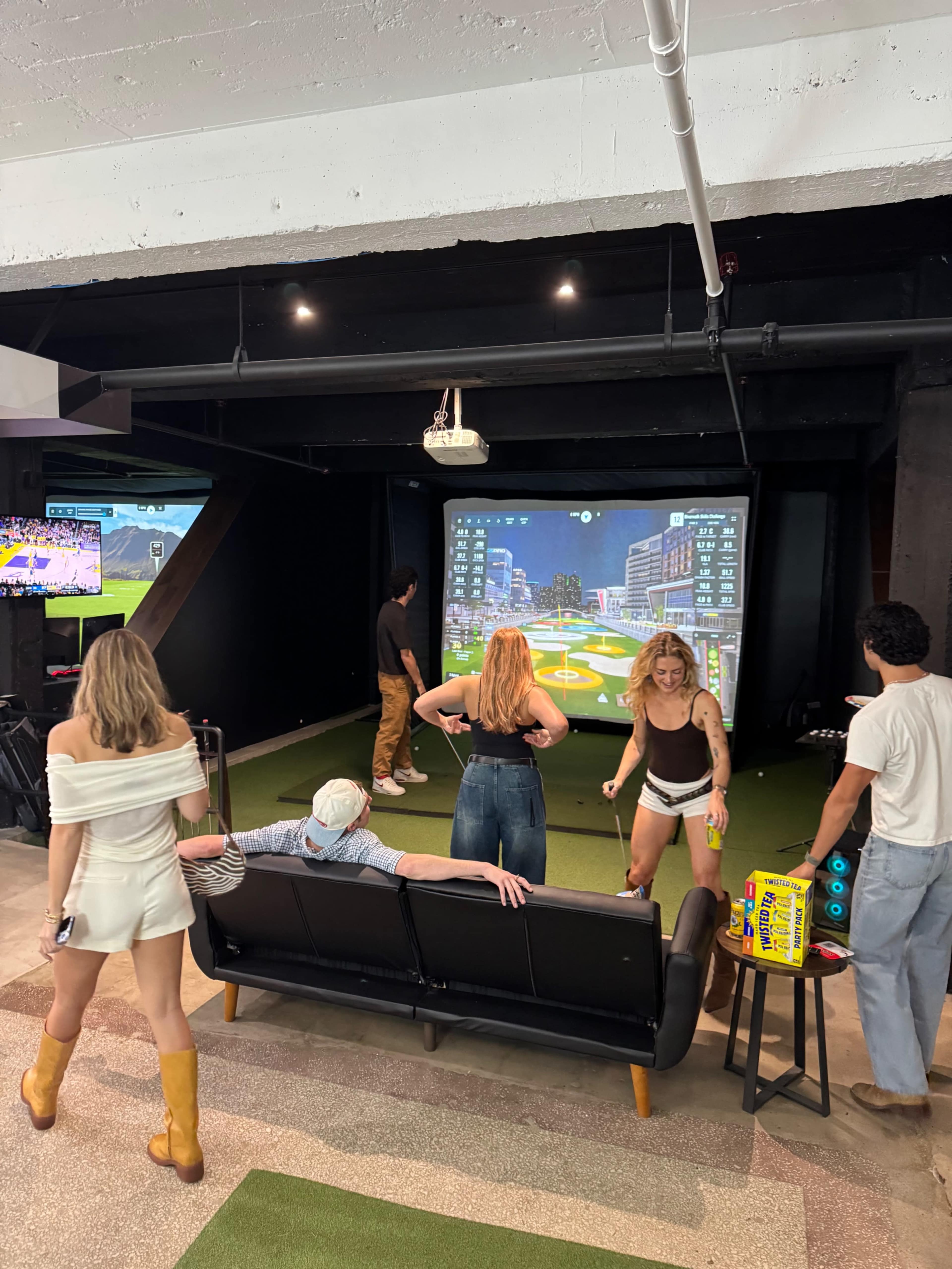 A group of five people is enjoying a golf simulation in a recreational space with a large projection screen and artificial turf.