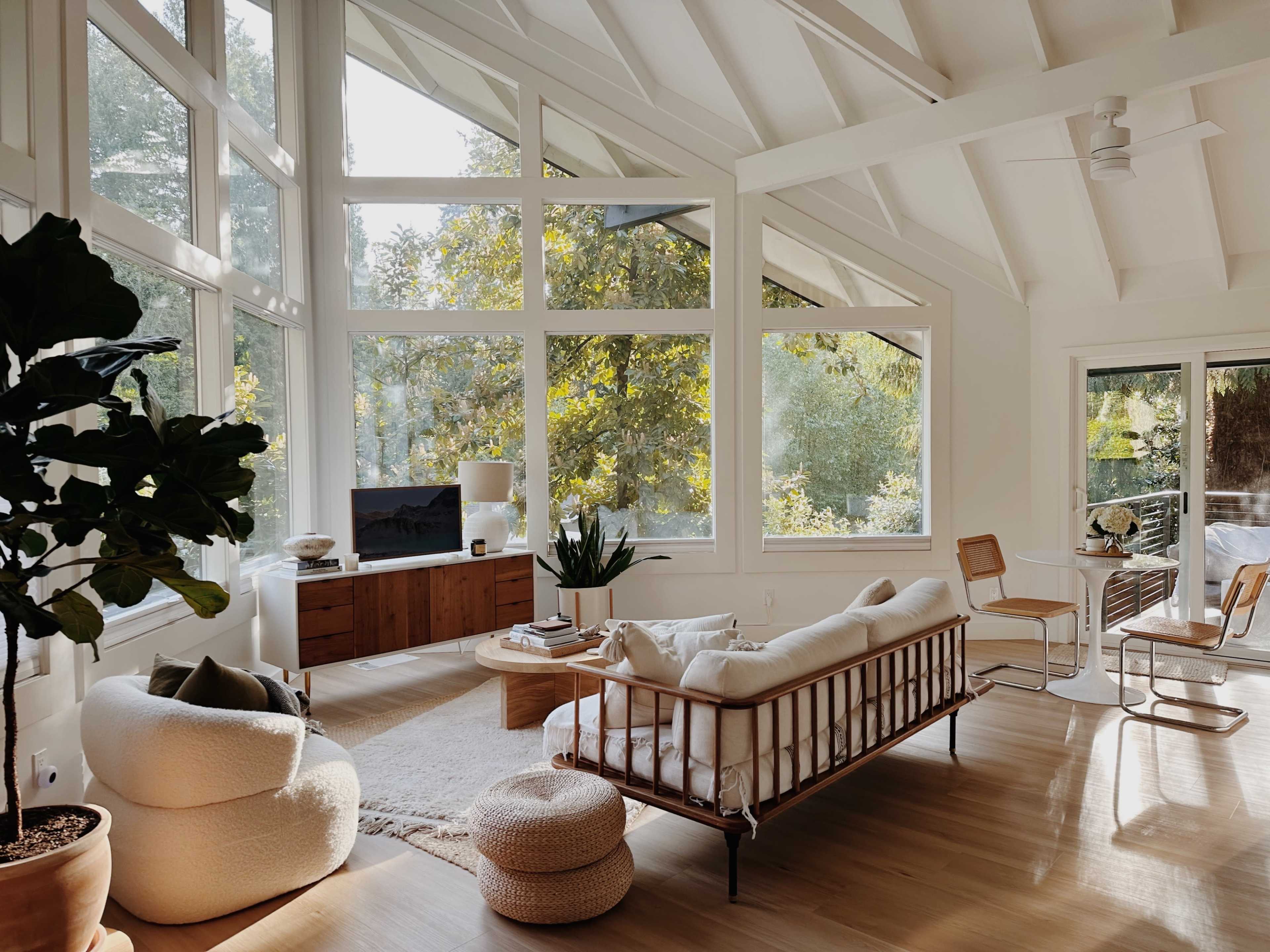 A bright living room features large windows, a white sofa, a wooden coffee table, and a television, surrounded by plants and natural light.