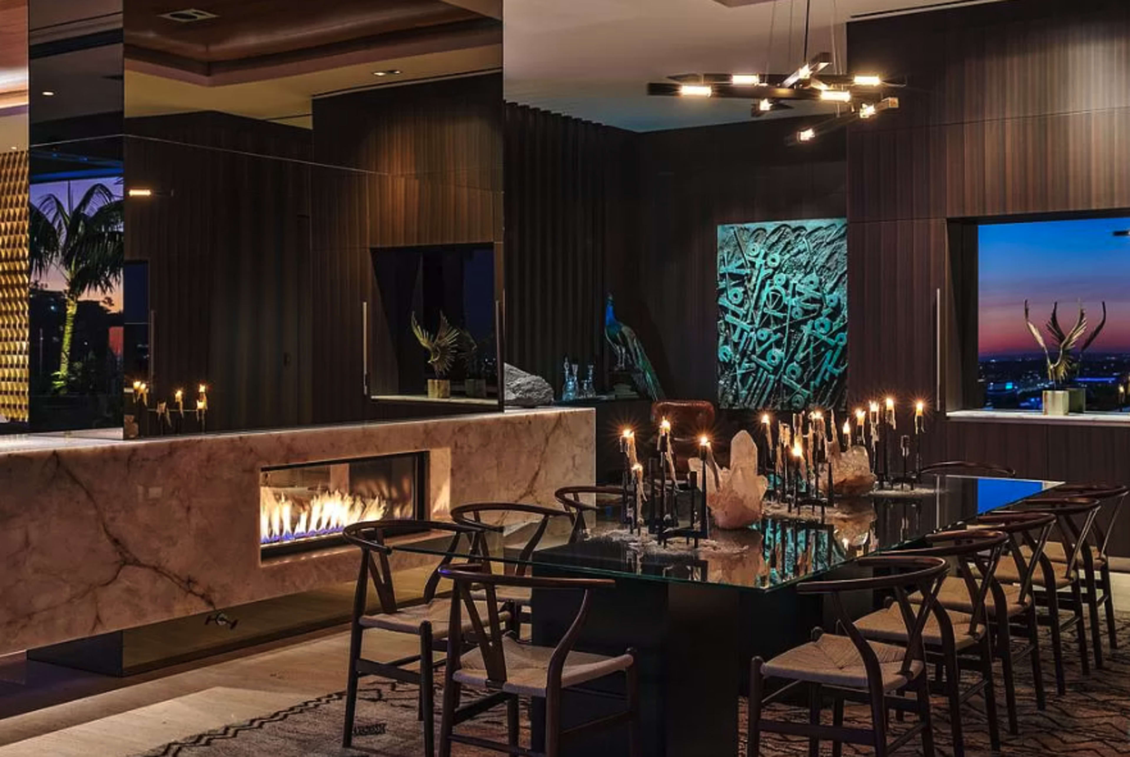 The image depicts a modern dining area with a large glass table surrounded by chairs, a fireplace integrated into the wall, and a panoramic view of the sunset through large windows.