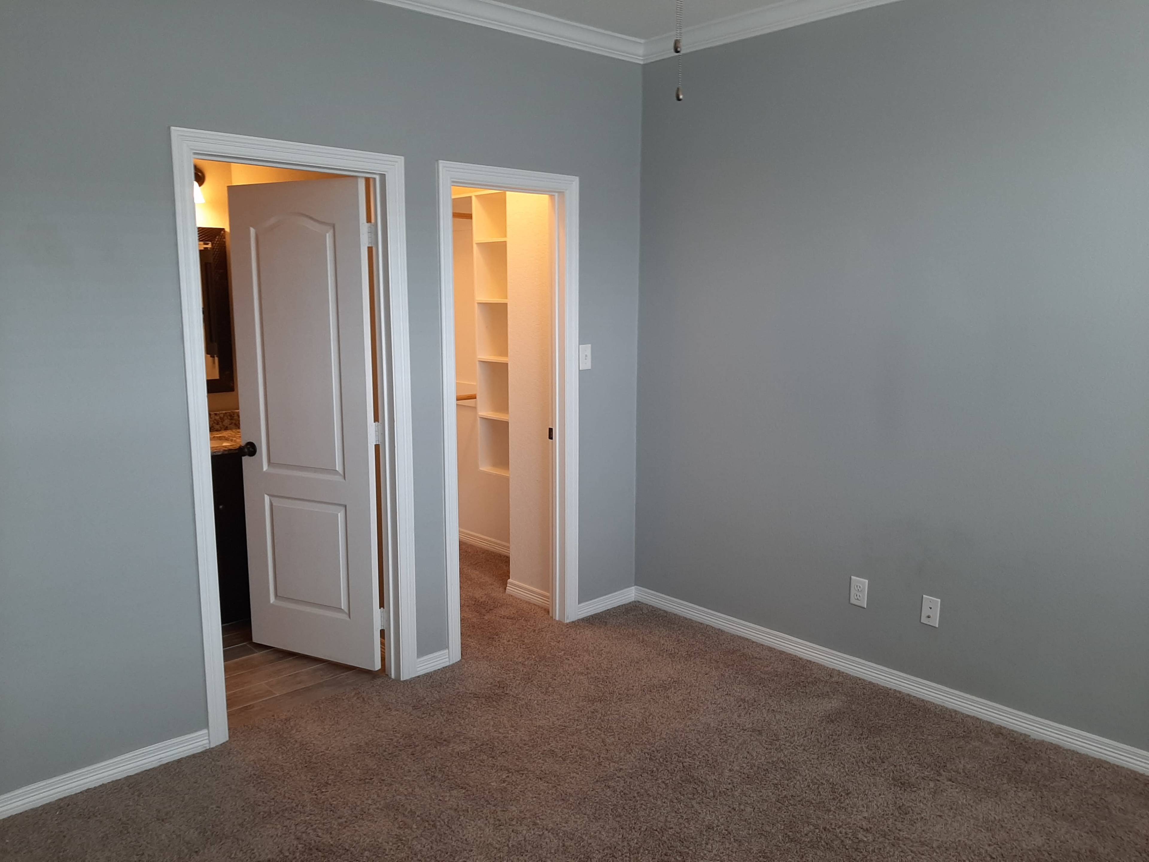 The image shows a sparsely furnished room with light gray walls, a carpeted floor, and two doorways leading to adjacent spaces.