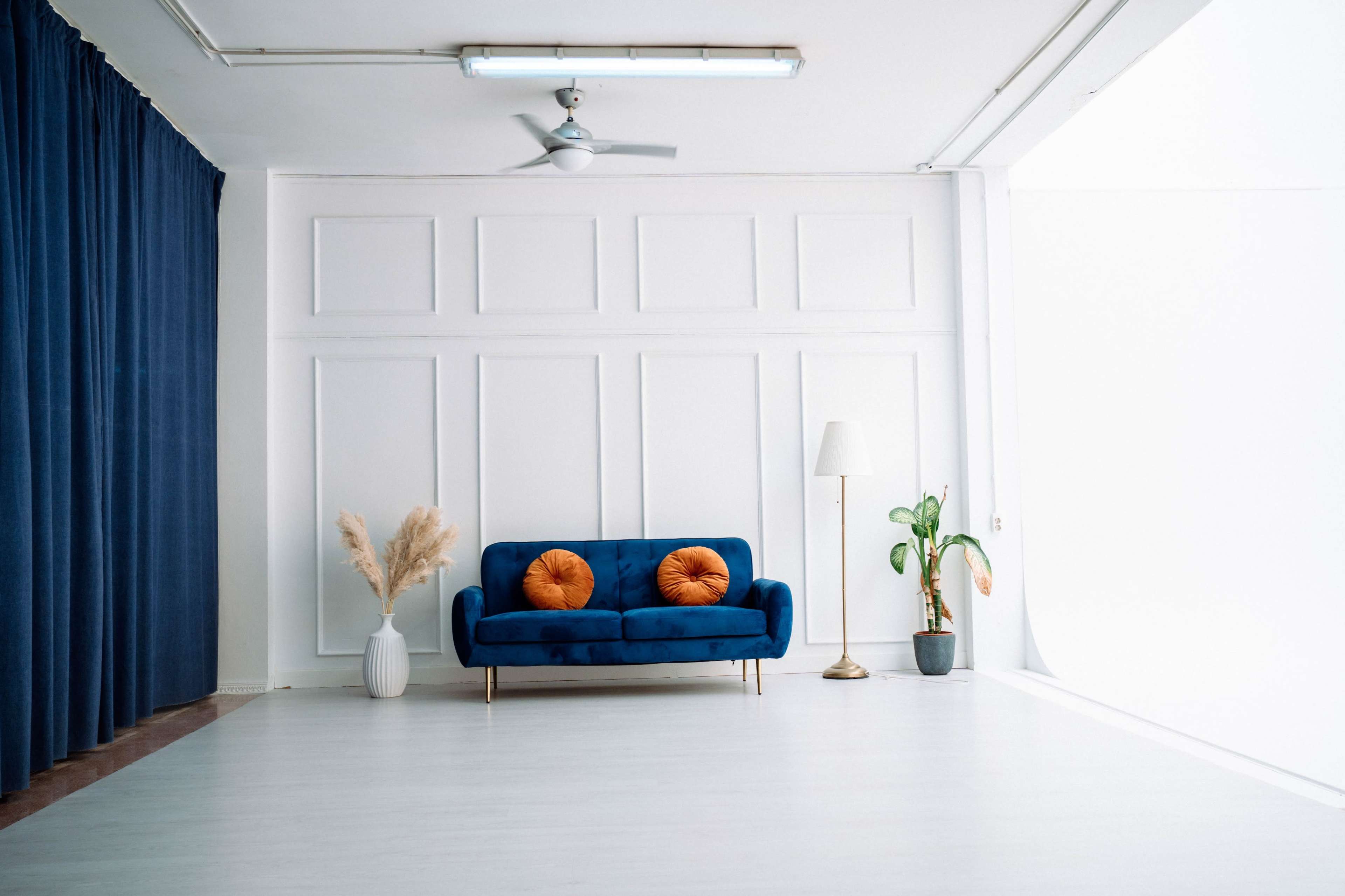 The image shows a bright room featuring a blue sofa with orange cushions, a floor lamp, and a potted plant, alongside a white wall and a blue curtain.