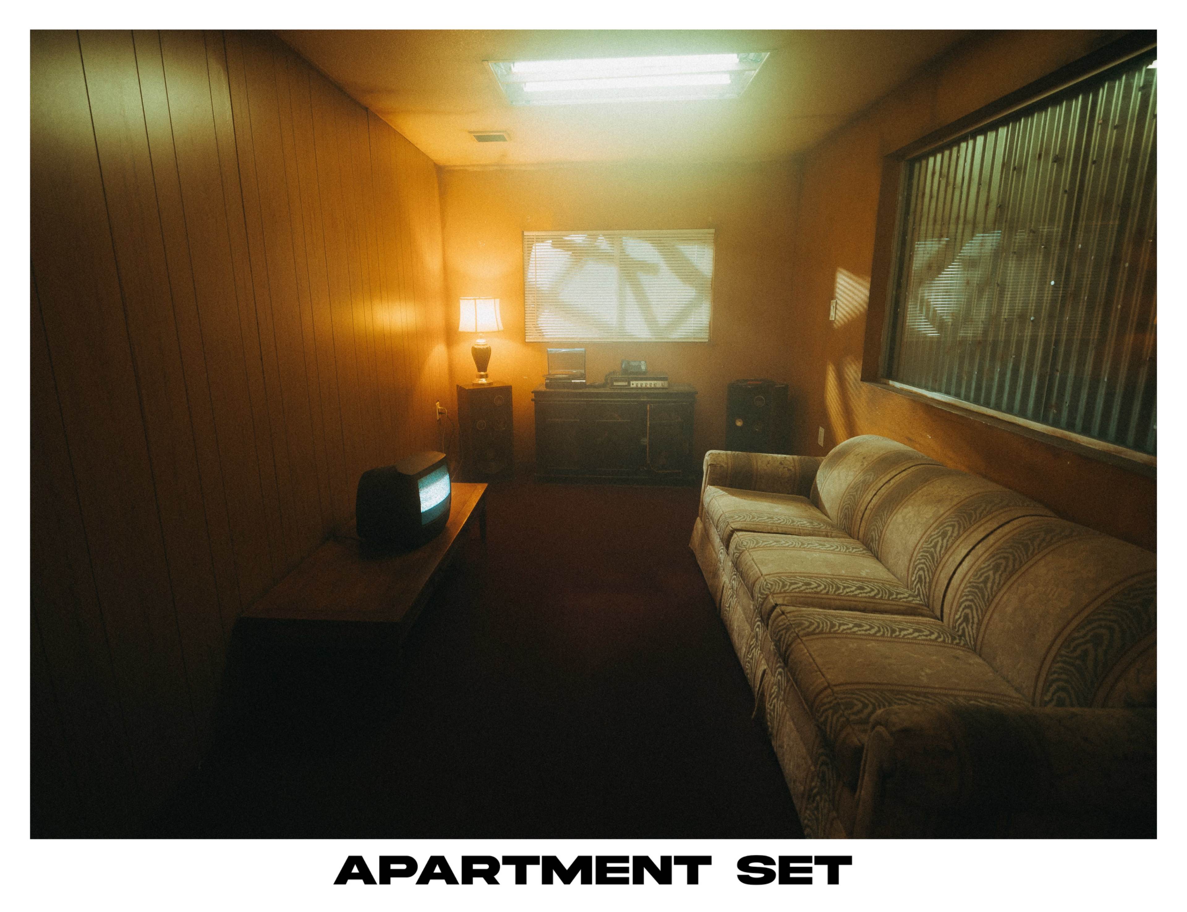 The image depicts a sparsely furnished apartment interior, featuring a sofa, a coffee table, a lamp, and an old television set.