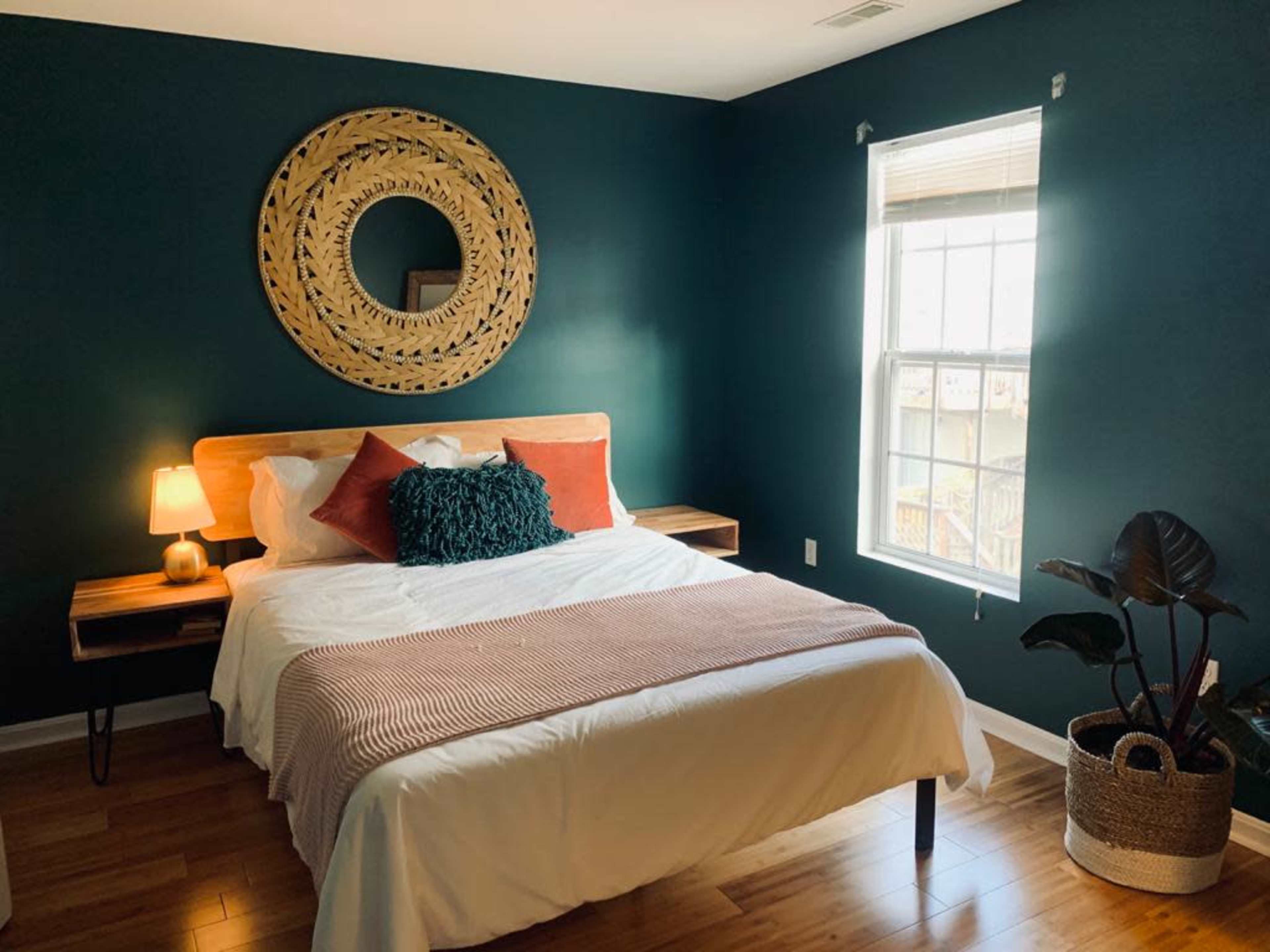 A neatly arranged bedroom featuring a bed with white linens, orange and teal pillows, a round decorative mirror on the wall, and a window allowing natural light.