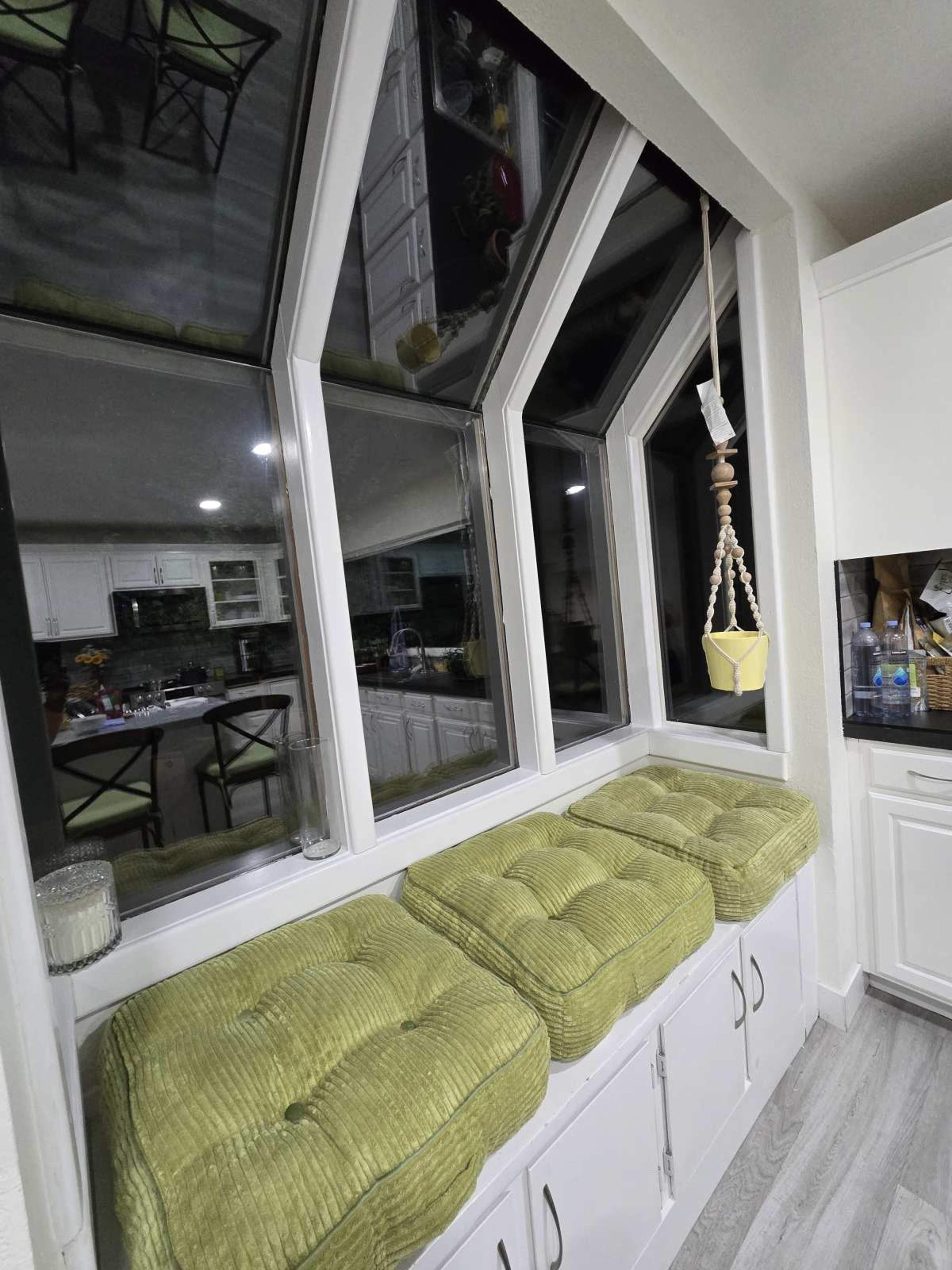 The image shows a kitchen nook with large angular windows, featuring three green cushioned seats on a white bench and a hanging planter.