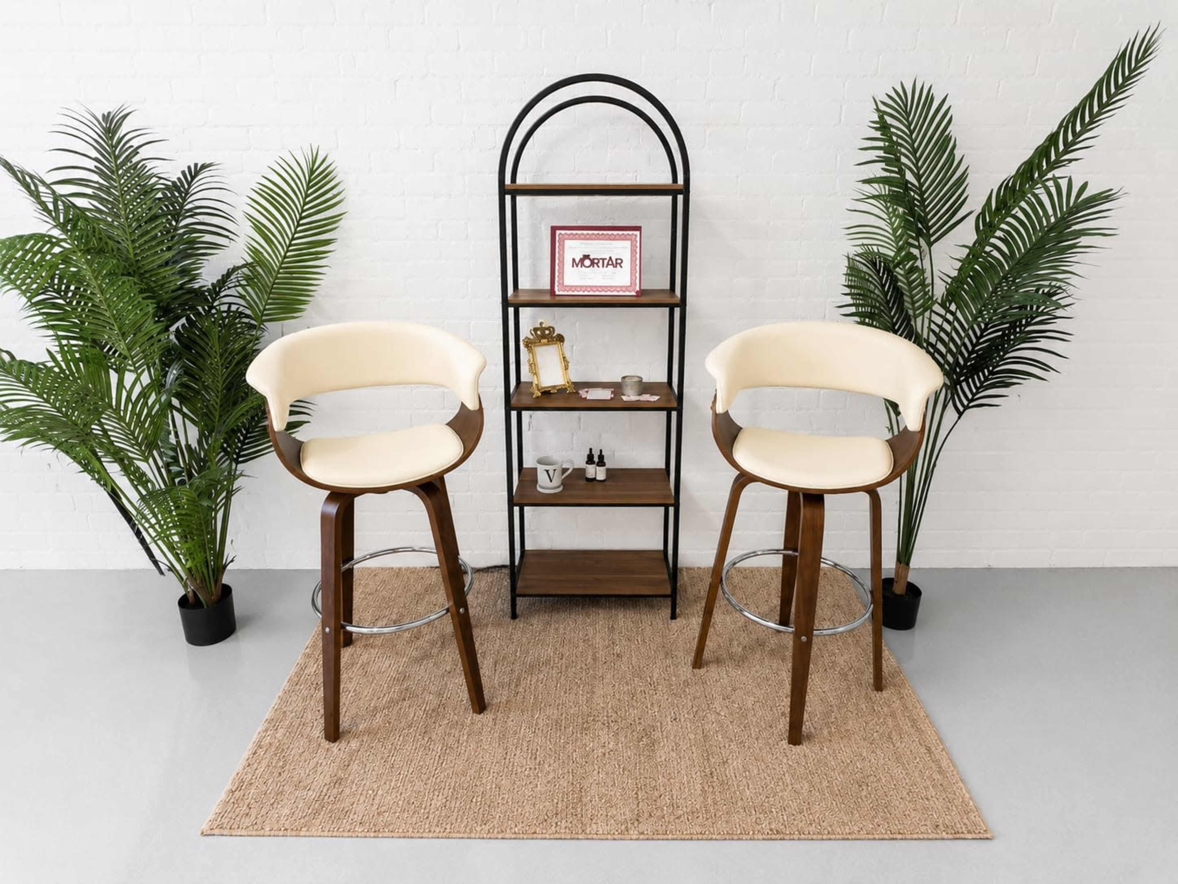 The image shows a minimalist setup with two cream-colored bar stools flanking a wooden shelving unit adorned with decorative items, surrounded by potted palm plants on a textured rug.