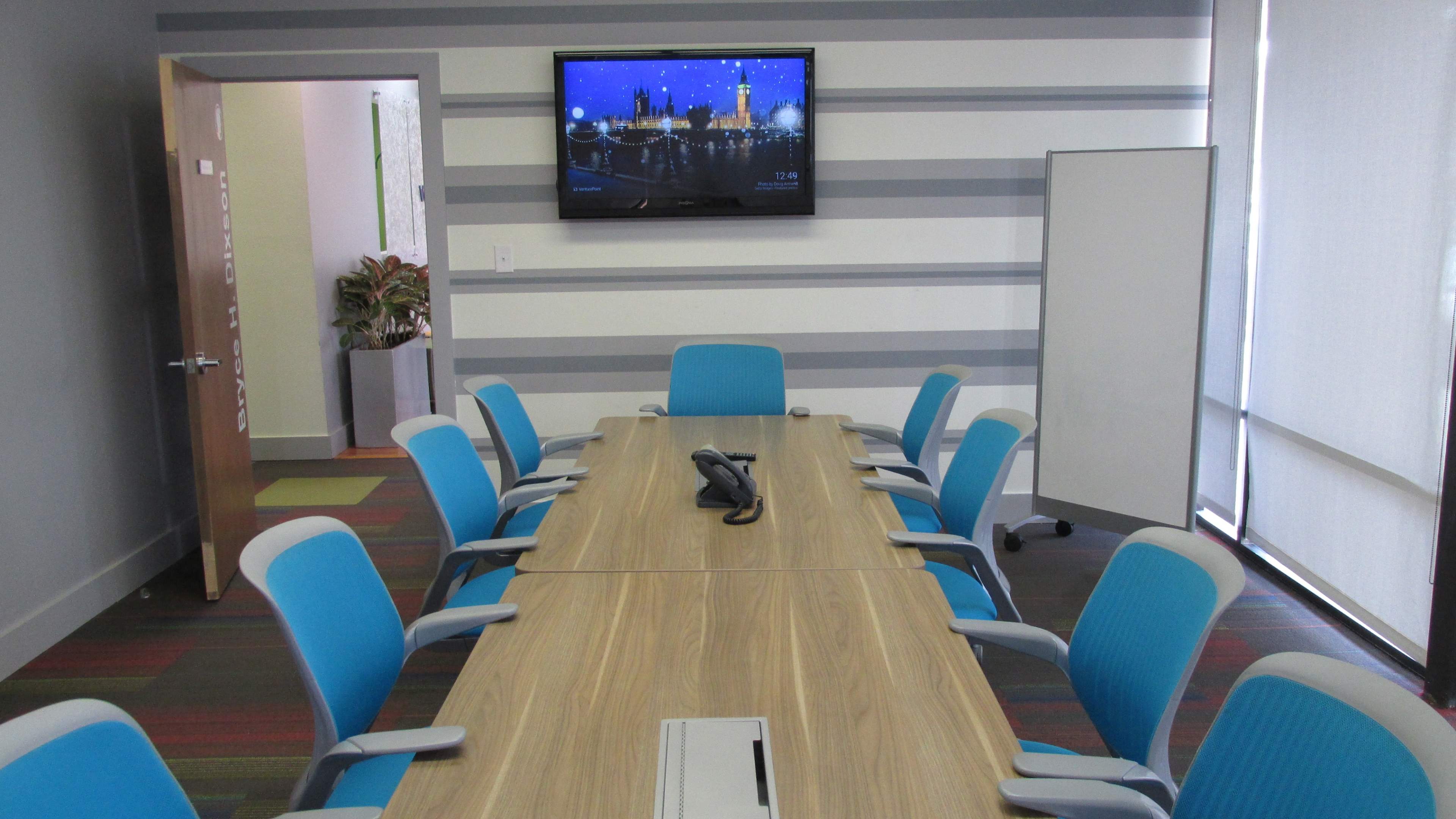 A modern conference room features a long wooden table surrounded by blue ergonomic chairs, with a wall-mounted screen displaying a cityscape in the background.