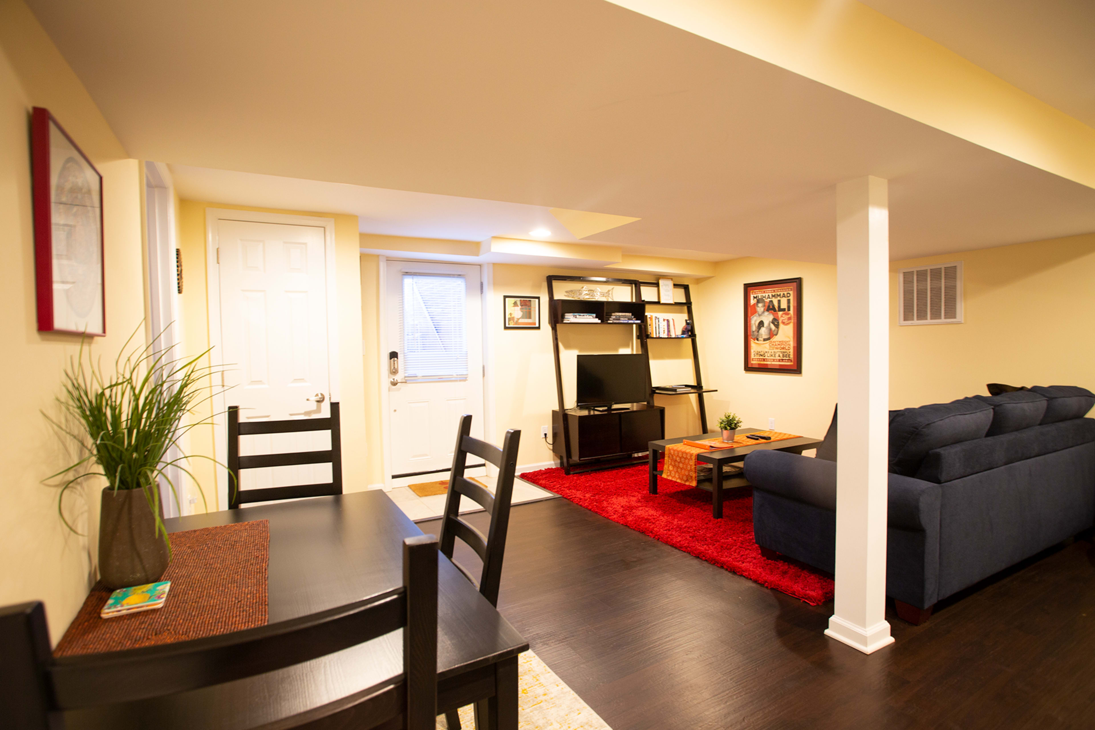 The image shows a well-lit basement living area with a dining table, a sofa, a media shelf, and a door leading outside.