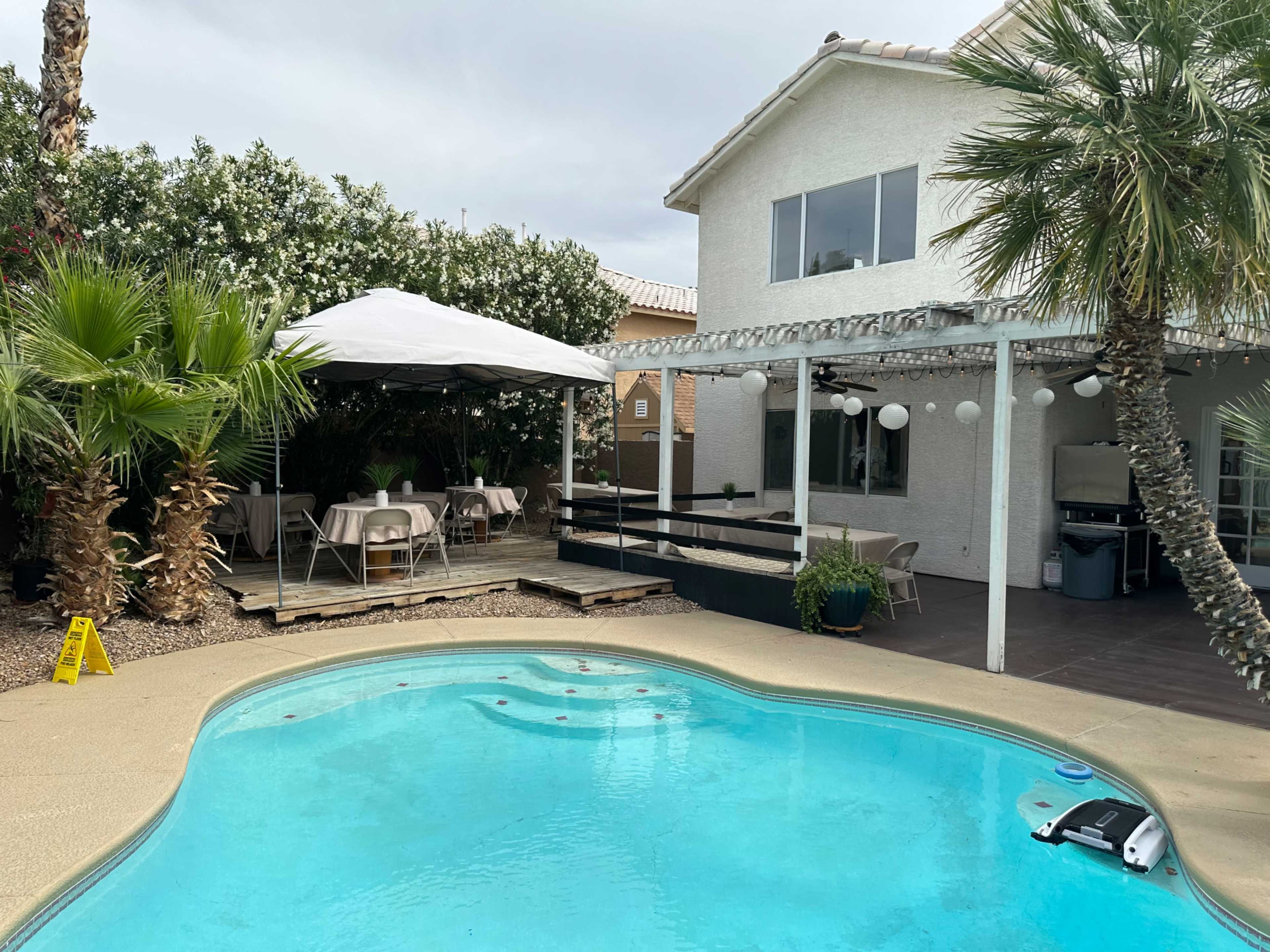 The image shows a backyard with a clear blue pool, a shaded patio area with tables, and a white house in the background.