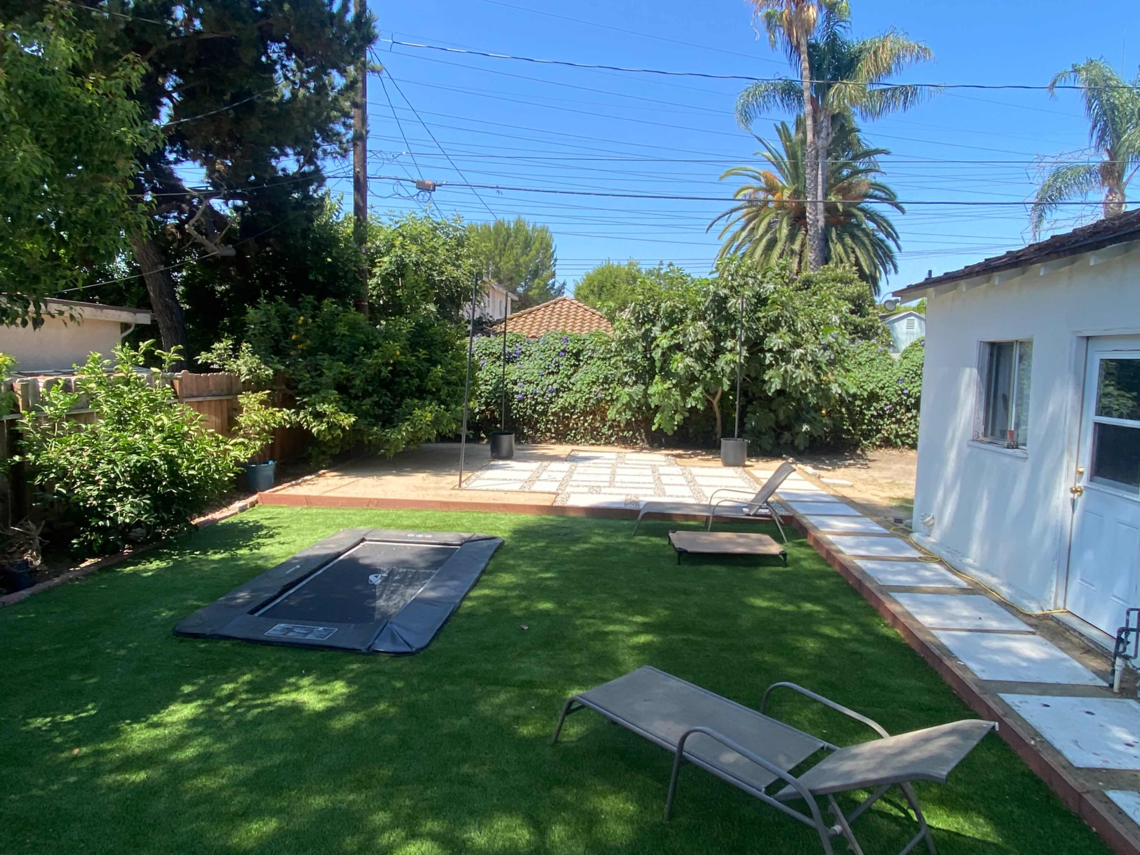 The image shows a private backyard with artificial turf, a lounge chair, a trampoline, and a paved area surrounded by trees and shrubs.