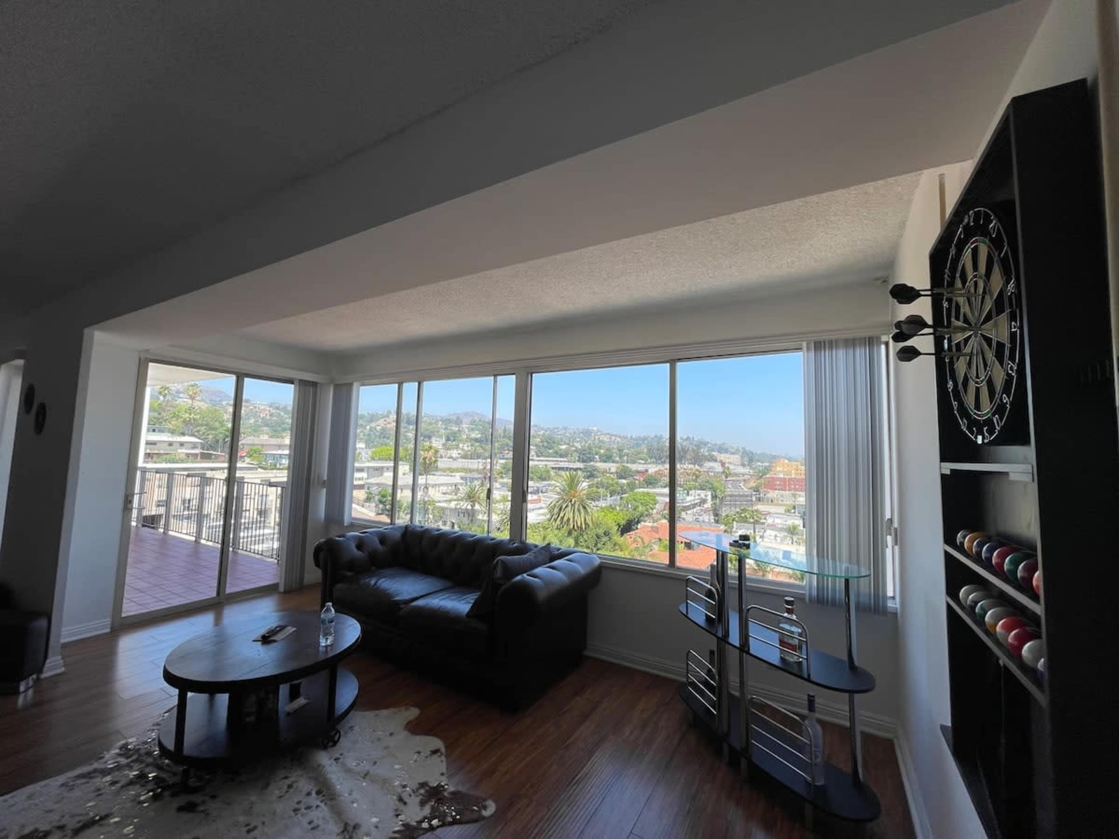 A modern living room features a leather couch, a round coffee table, and large windows overlooking a hillside cityscape.