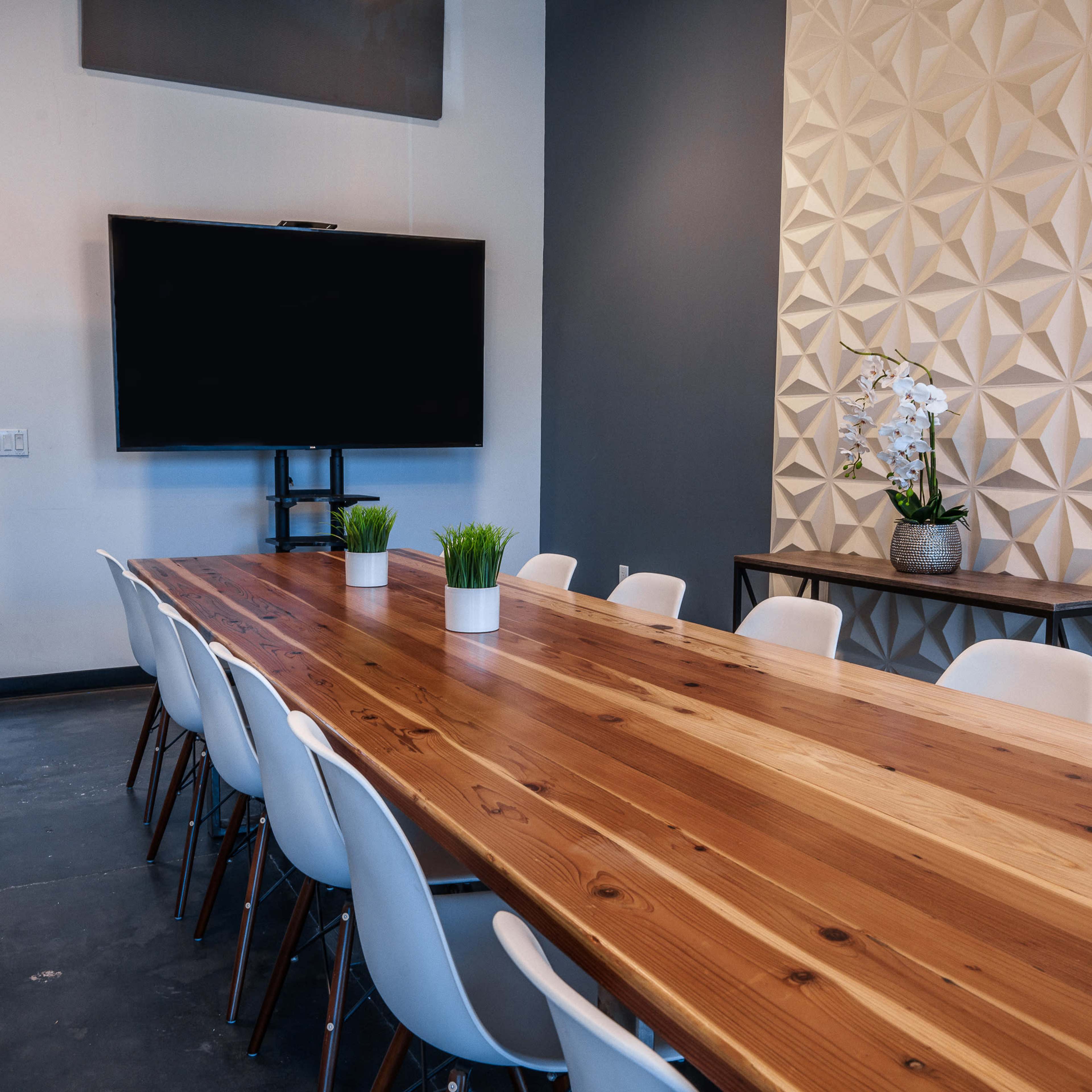 A long wooden conference table is set with white chairs, and there are two plants on the table beside a television mounted on the wall and a textured decorative panel.
