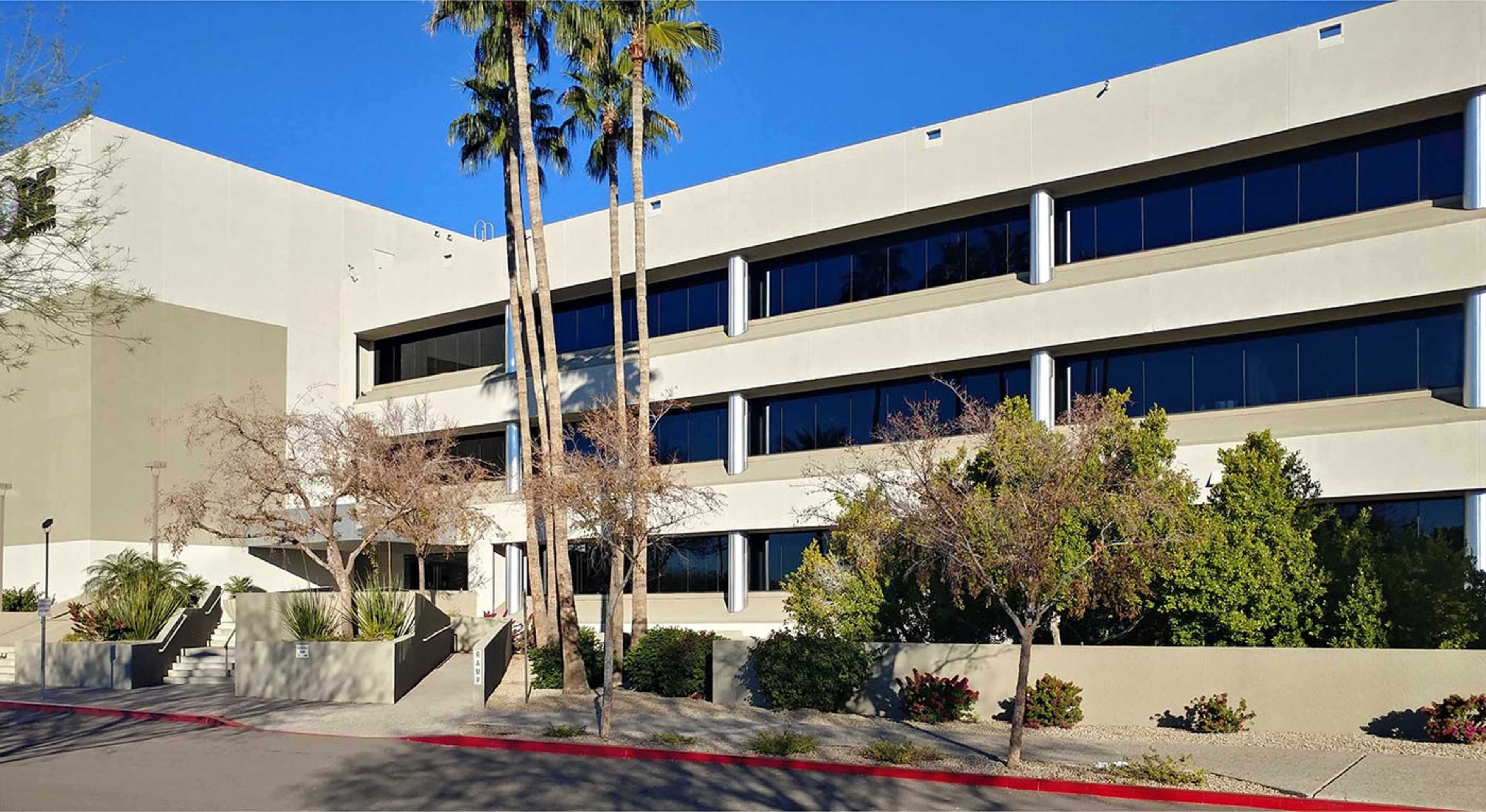 A modern three-story commercial building with large windows and surrounding palm trees and shrubs.