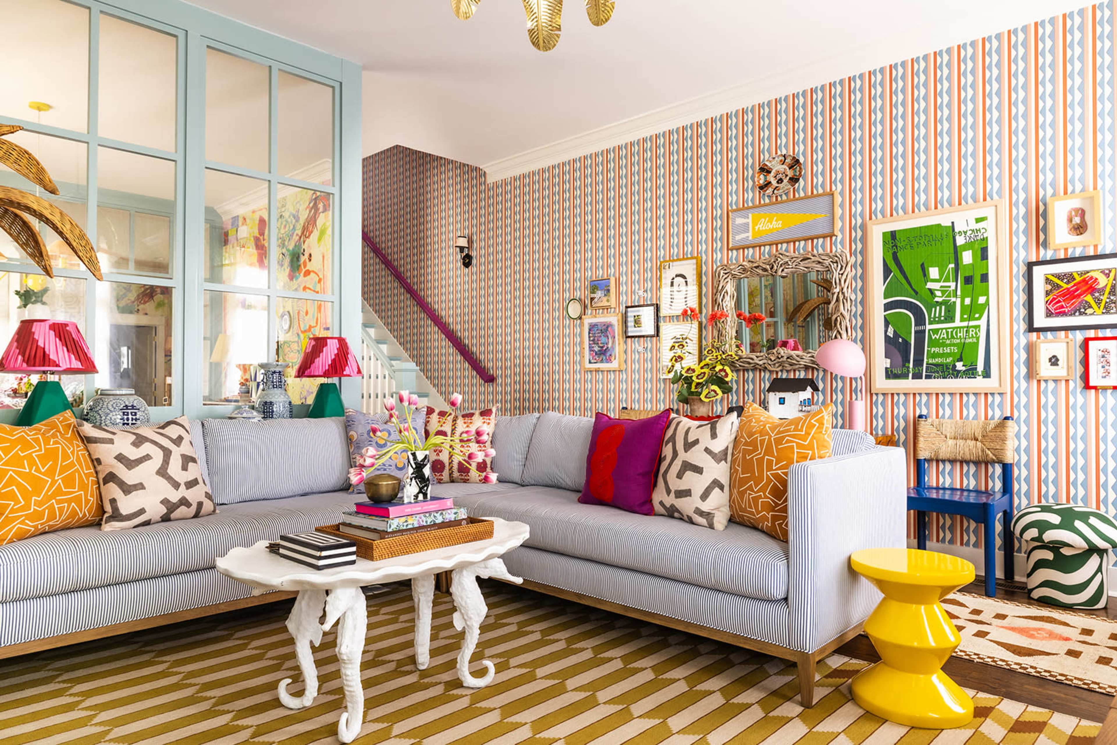 The living room features a light blue sectional sofa adorned with colorful patterned pillows, a coffee table with decorative items, and vibrant wallpaper with geometric designs.