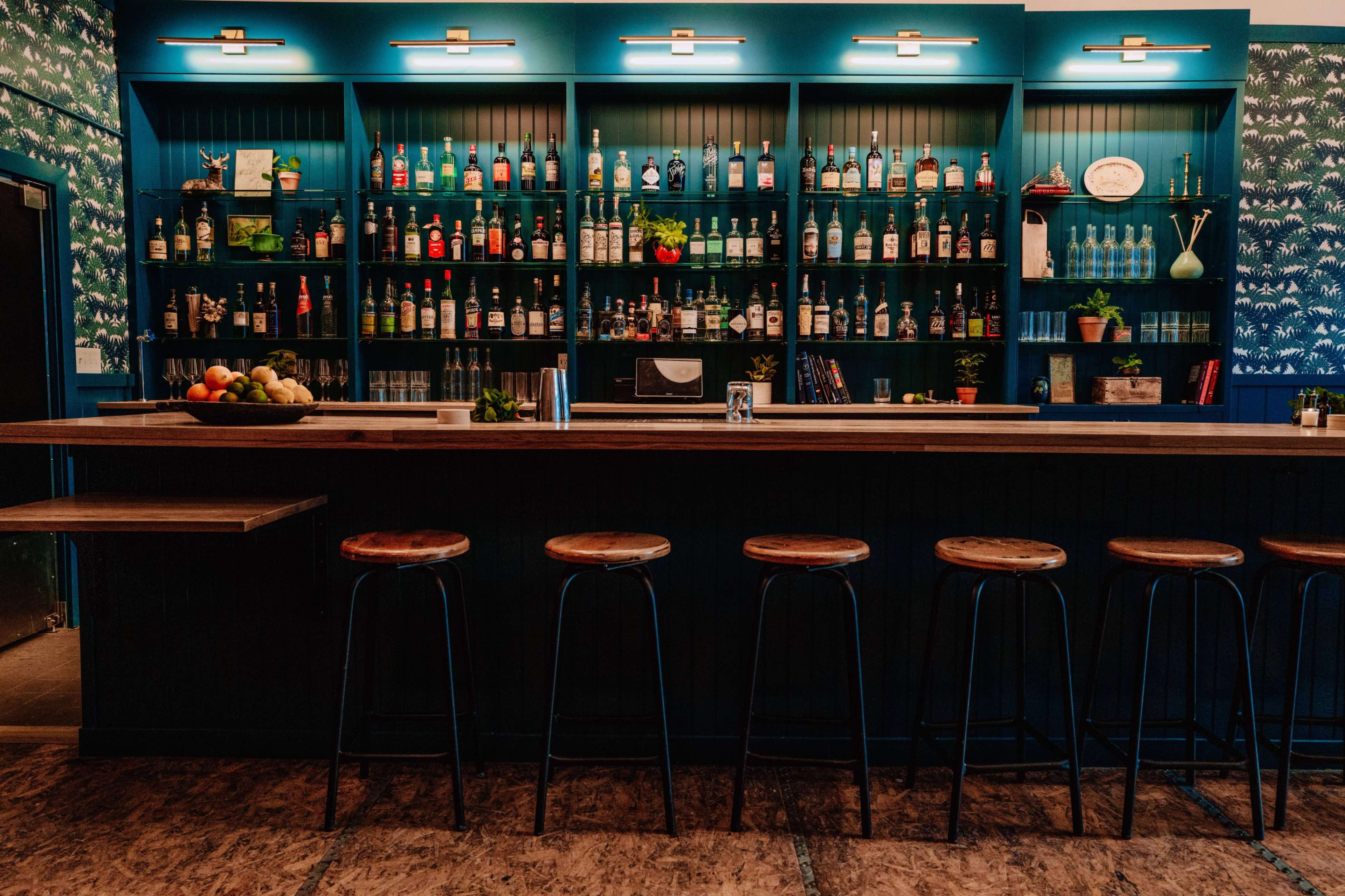 A bar with a wooden countertop and several metallic stools lined up in front of a well-stocked back shelf of bottles and glassware.