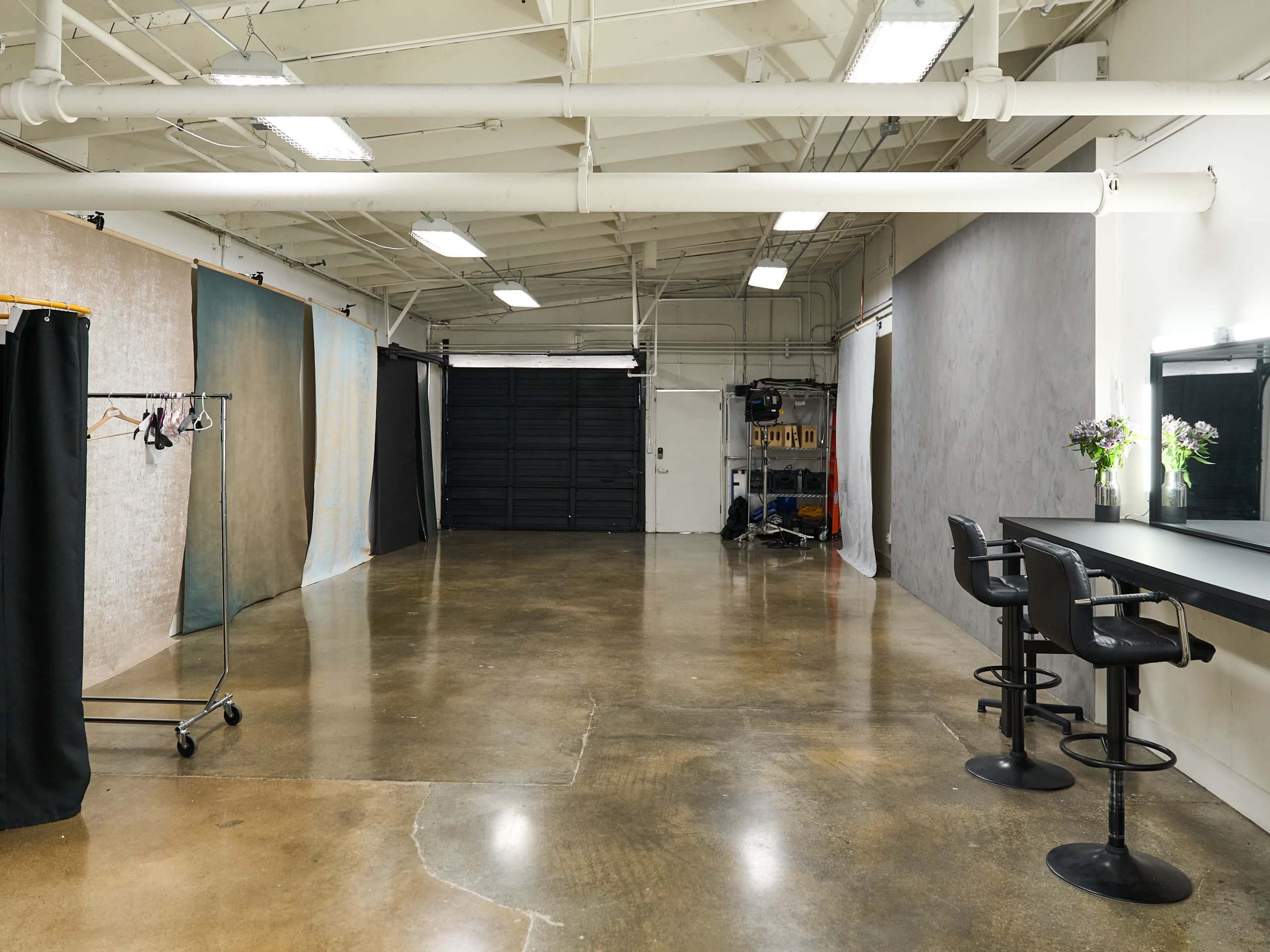 An empty photography studio with a concrete floor, a backdrop setup, and a mirrored vanity area with stools.