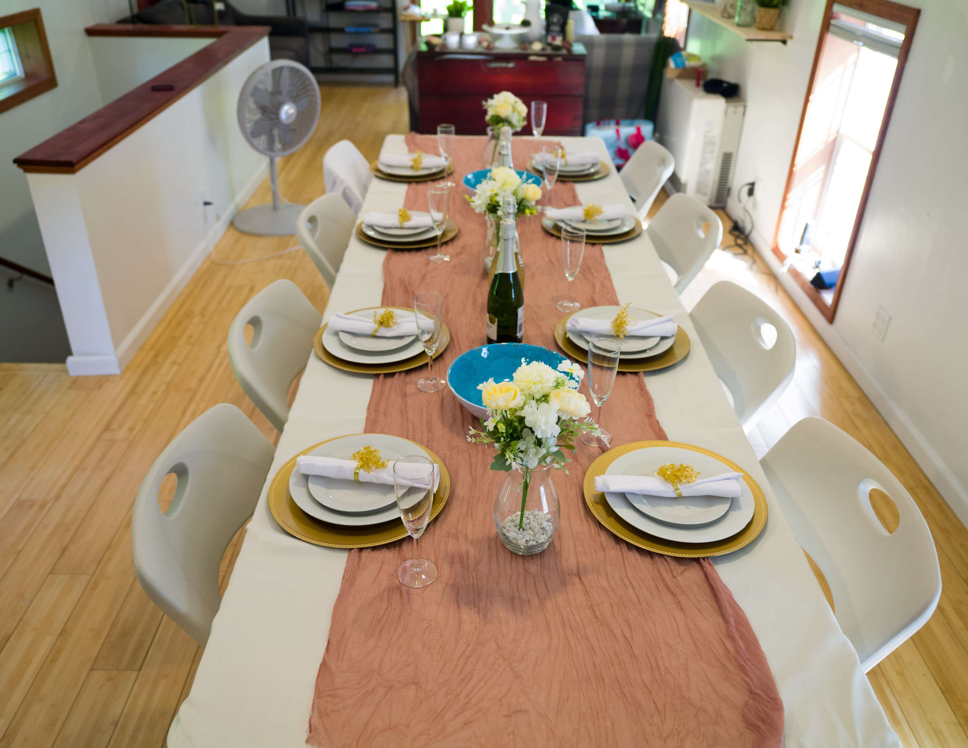 A long dining table is set with white plates, golden chargers, glassware, and floral centerpieces, against a backdrop of a bright, airy room.