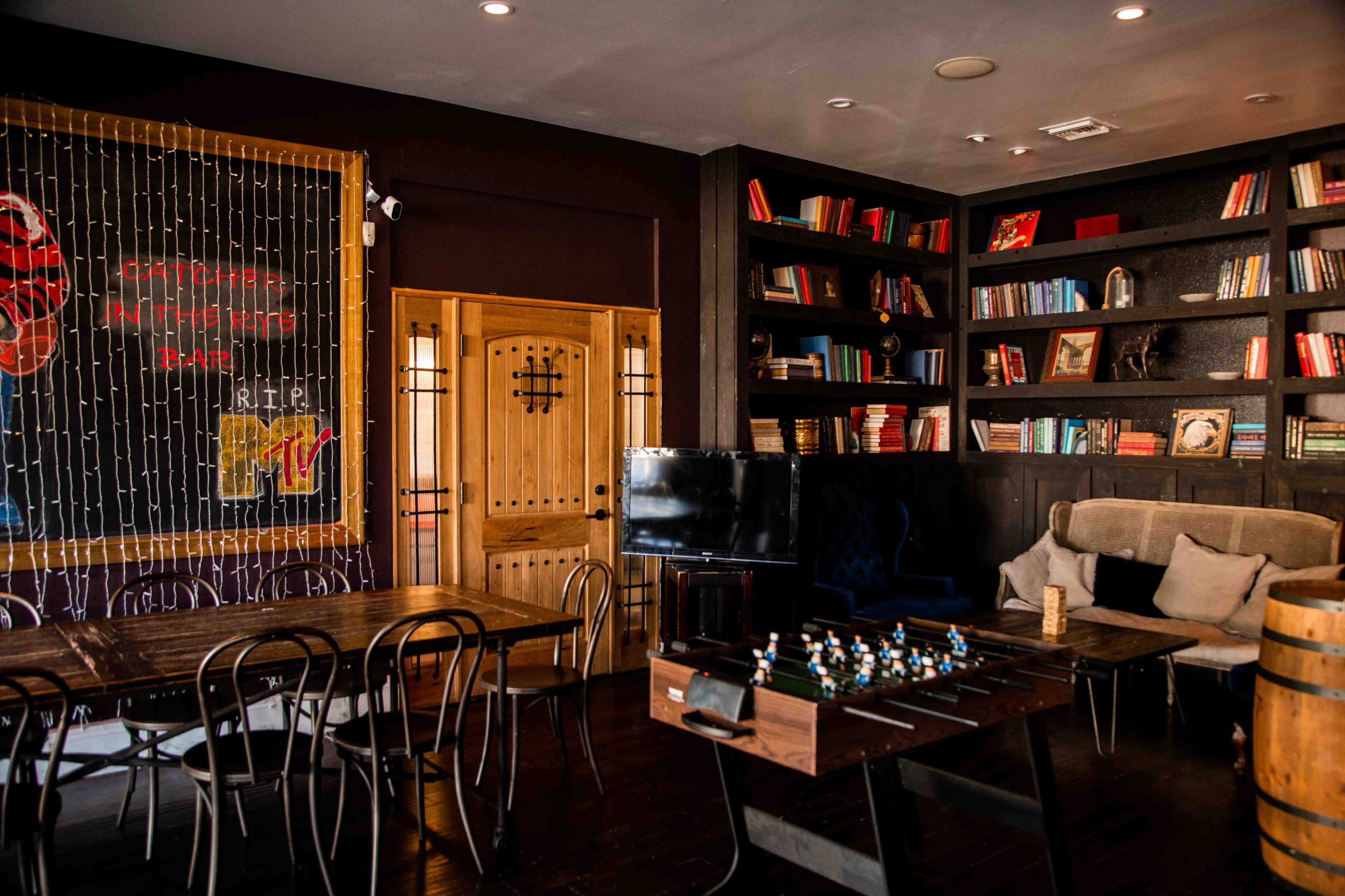 The image shows a cozy indoor space furnished with wooden tables, a foosball table, bookshelves filled with books, and a large television.