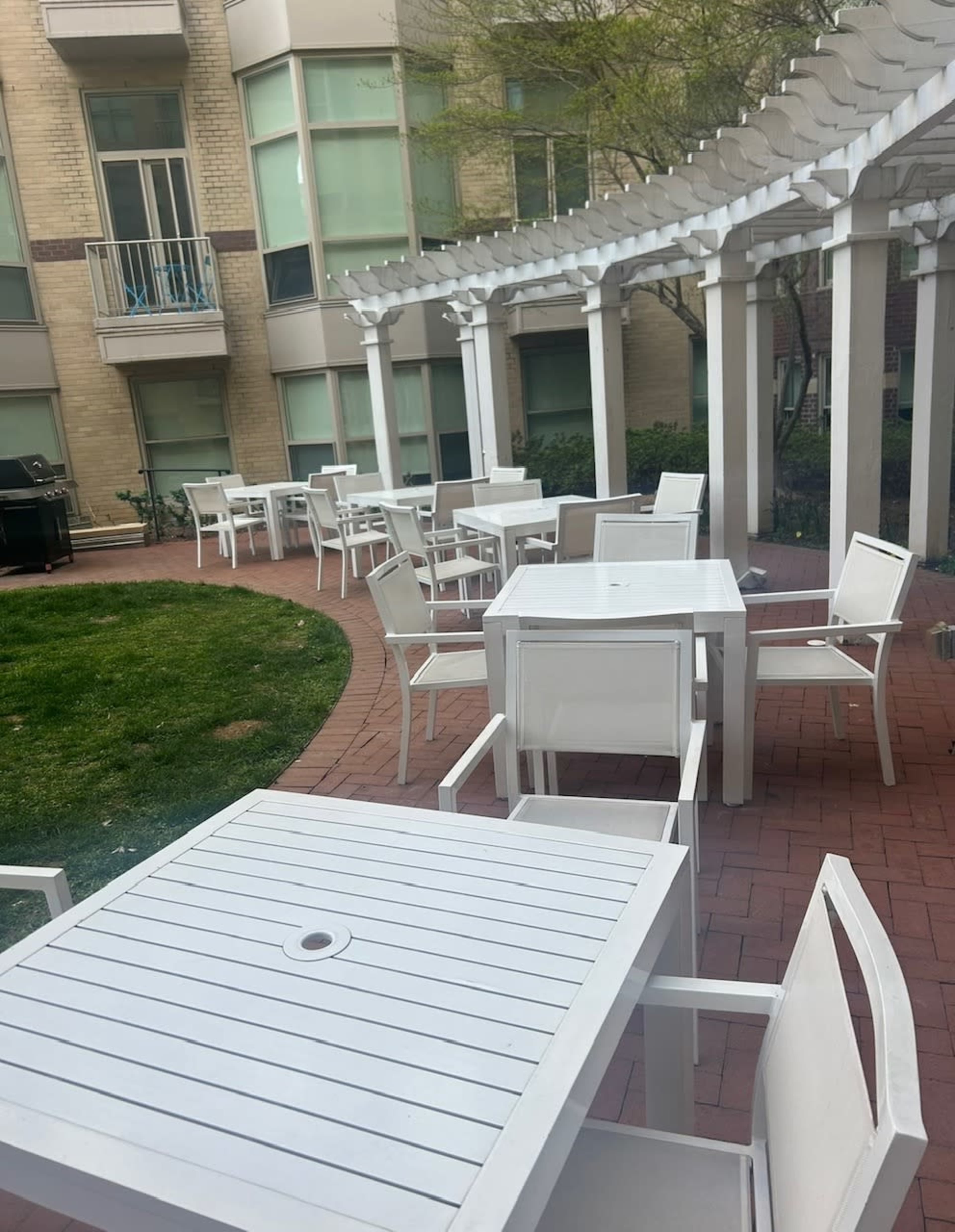 The image shows a courtyard with several white outdoor tables and chairs arranged on brick pathways surrounded by greenery and a white pergola.