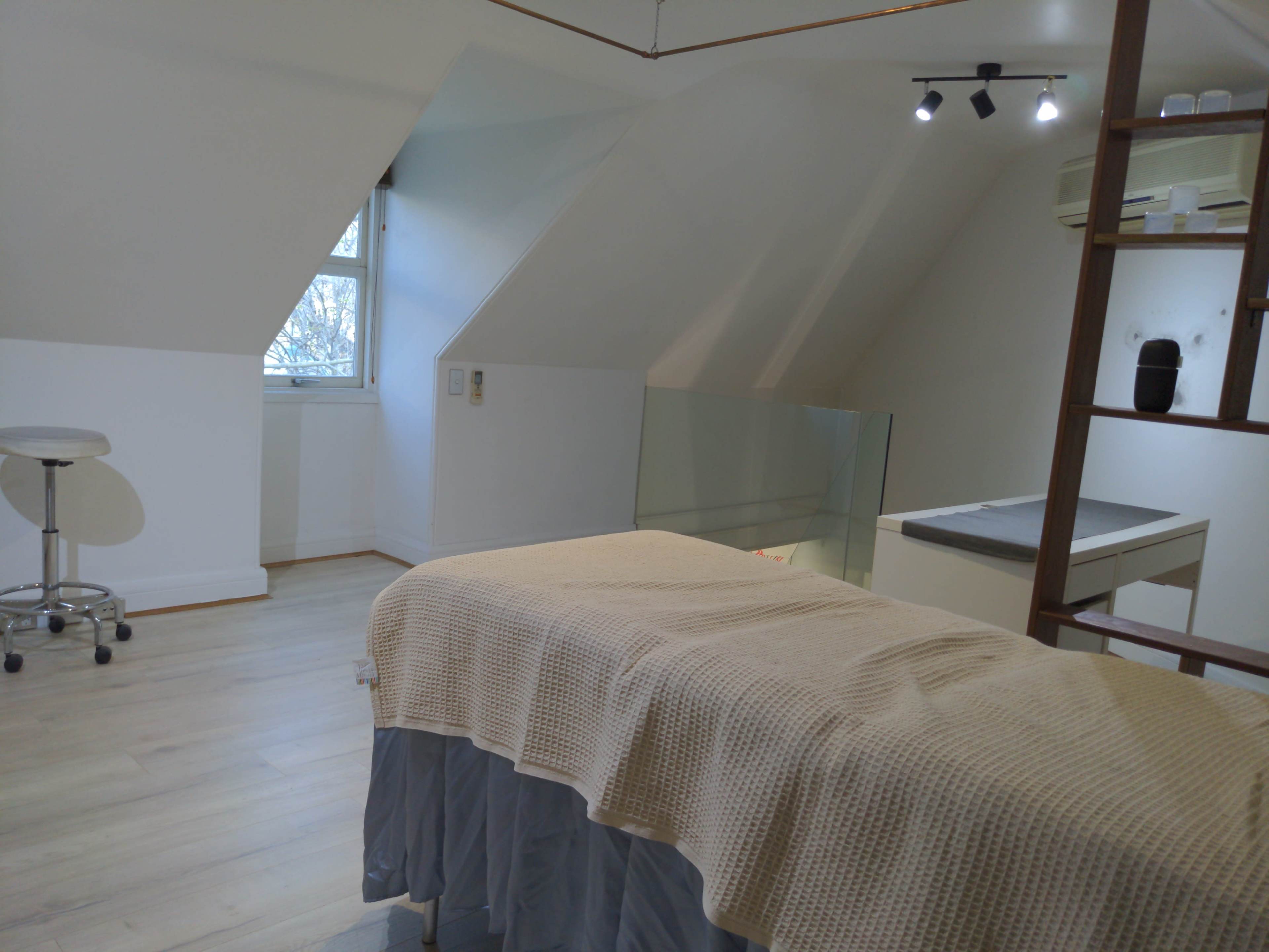 The image shows a clean, well-lit room with a treatment table covered in a beige cloth, a stool, and a work surface against a slanted wall.