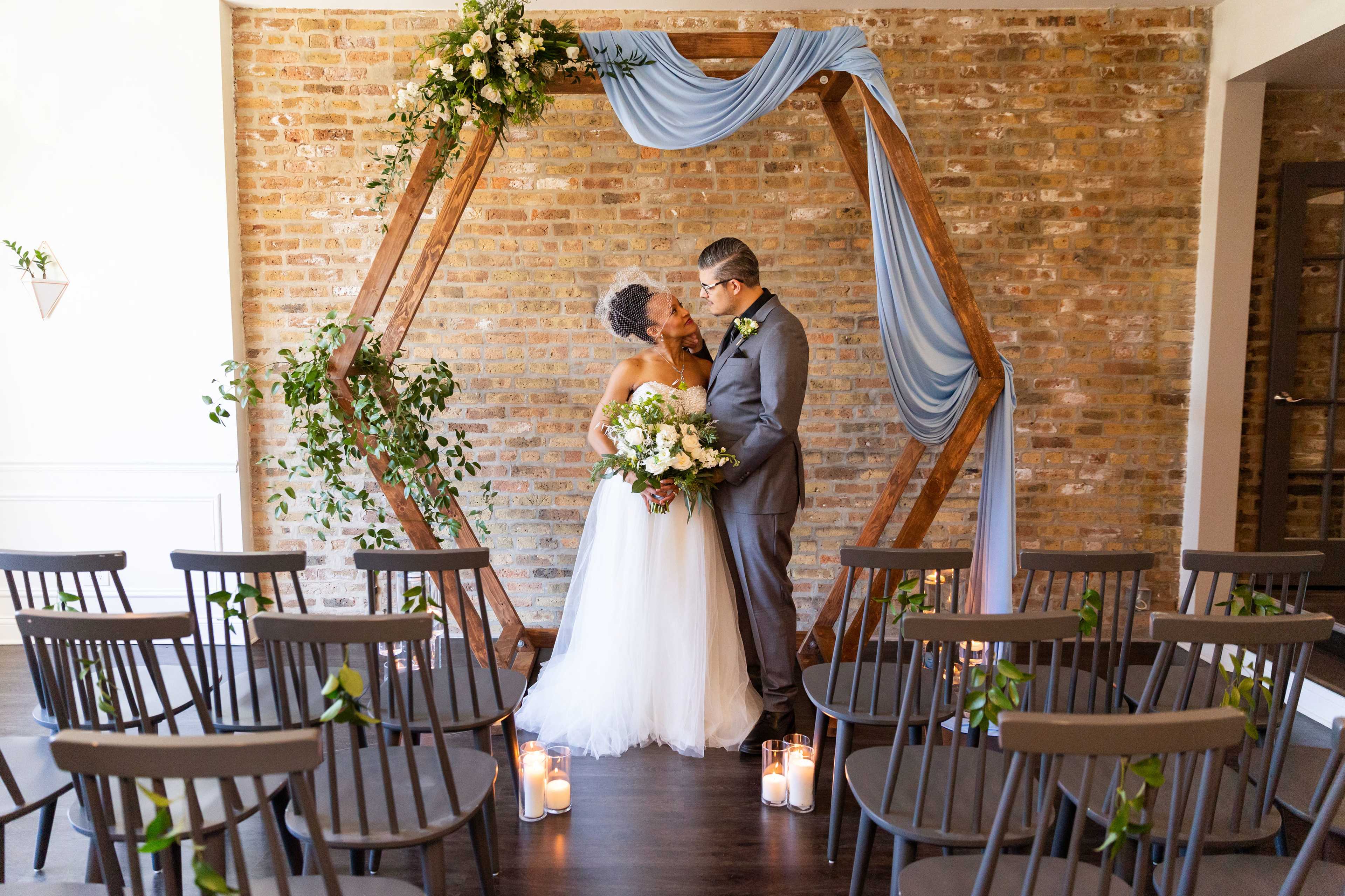 A bride and groom kiss under a floral arch in a brick-walled venue with chairs arranged for a wedding ceremony.