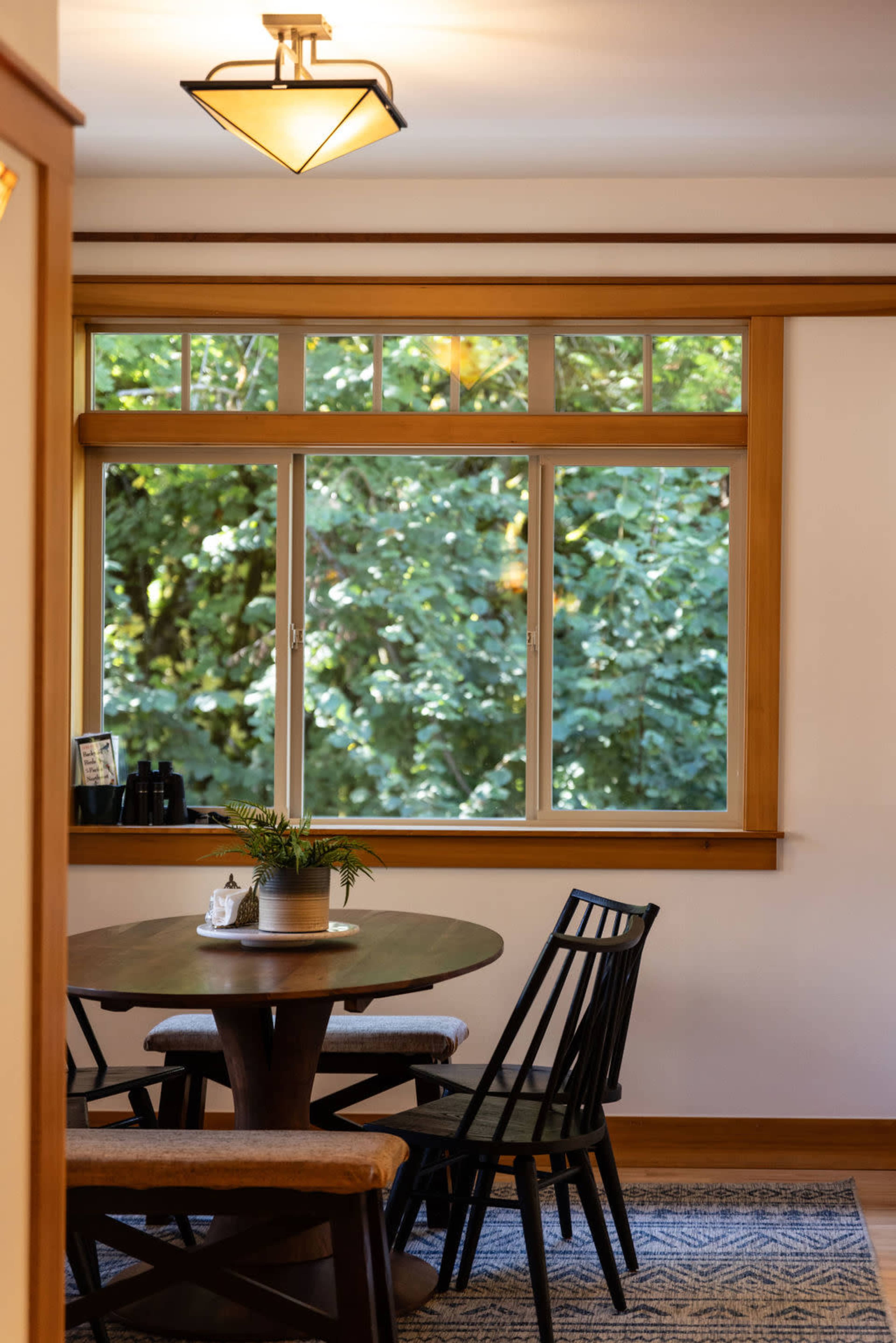 A round table with black chairs sits beneath a light fixture in a room with a large window overlooking greenery.