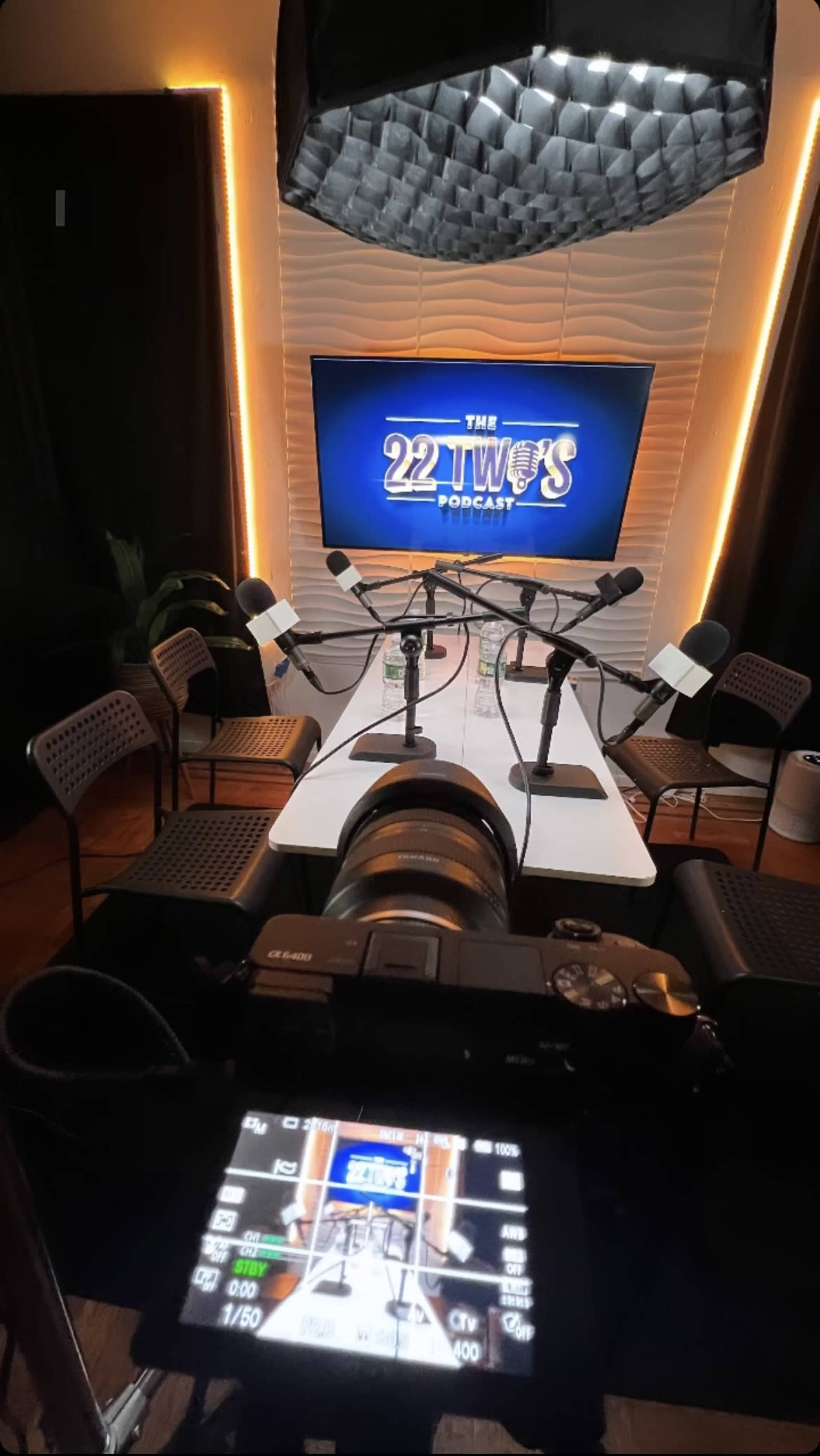 A camera is positioned in front of a podcast recording setup, featuring a table with microphones and chairs, illuminated by soft lighting and displaying a screen with the podcast title.