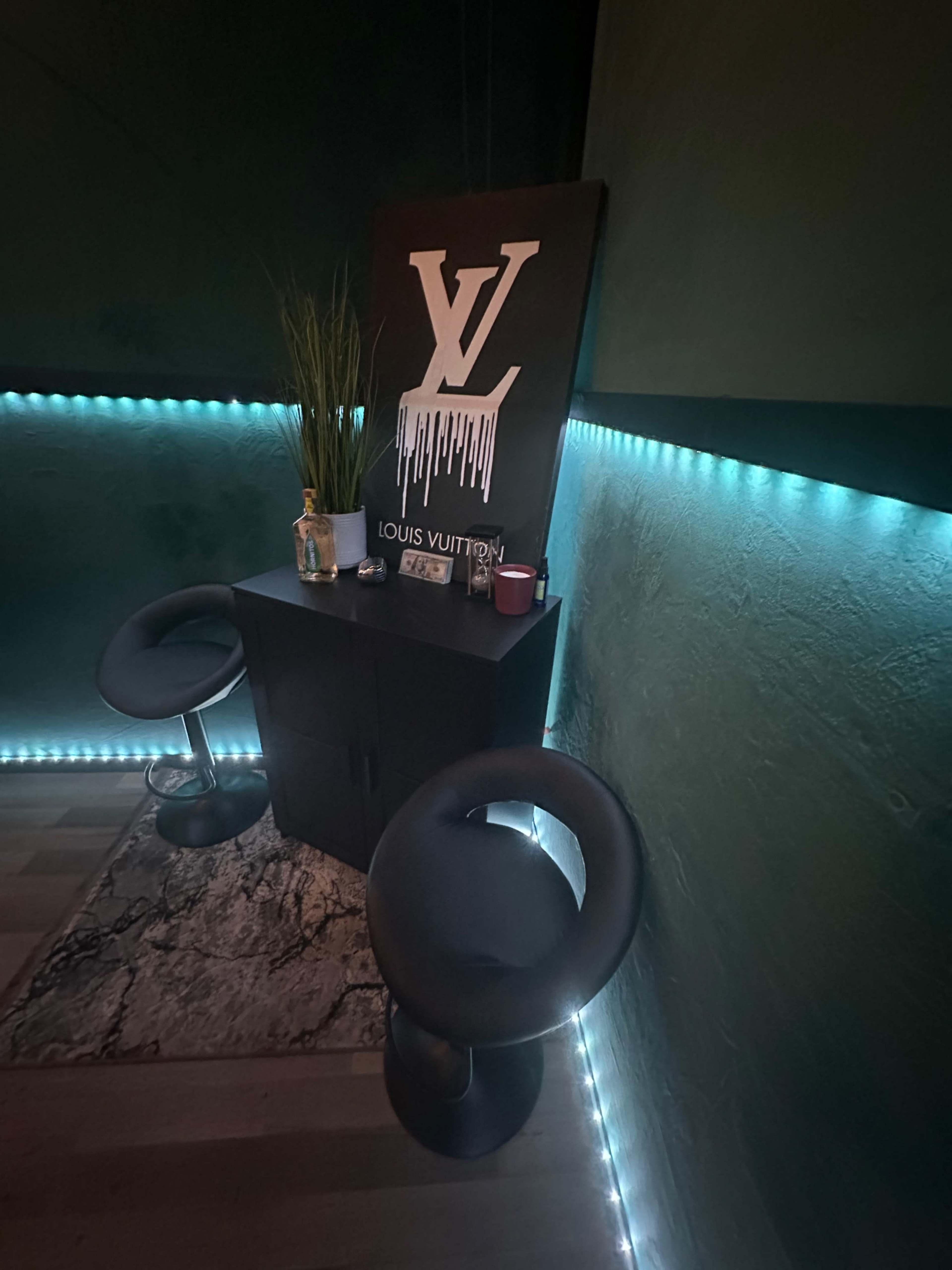 A corner of a room featuring two black stools, a black cabinet, and a large sign displaying the Louis Vuitton logo, illuminated by blue LED lights.