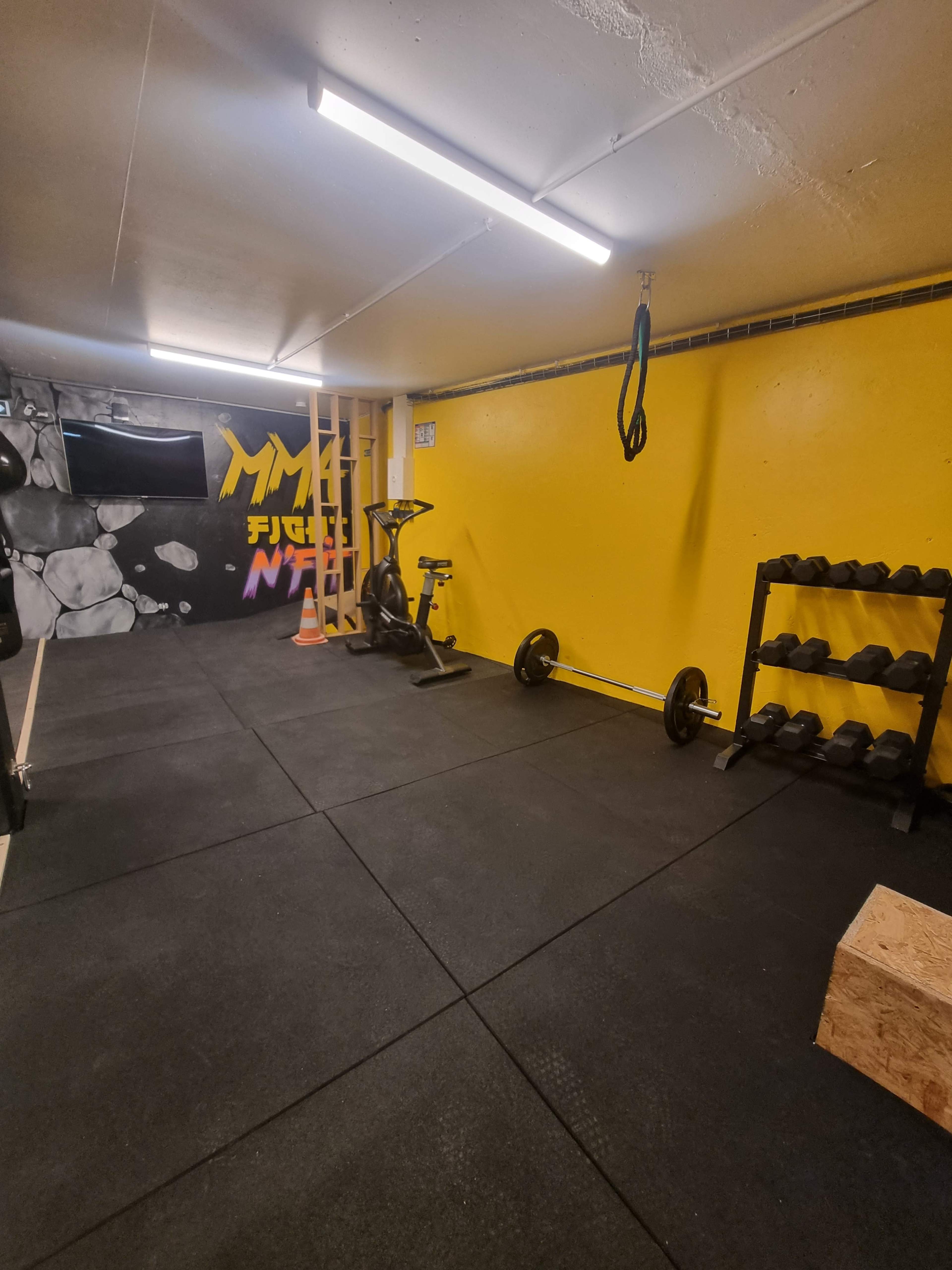 A small gym space with black rubber flooring, a yellow wall, a stationary bike, a set of dumbbells on a rack, and a wooden plyometric box.