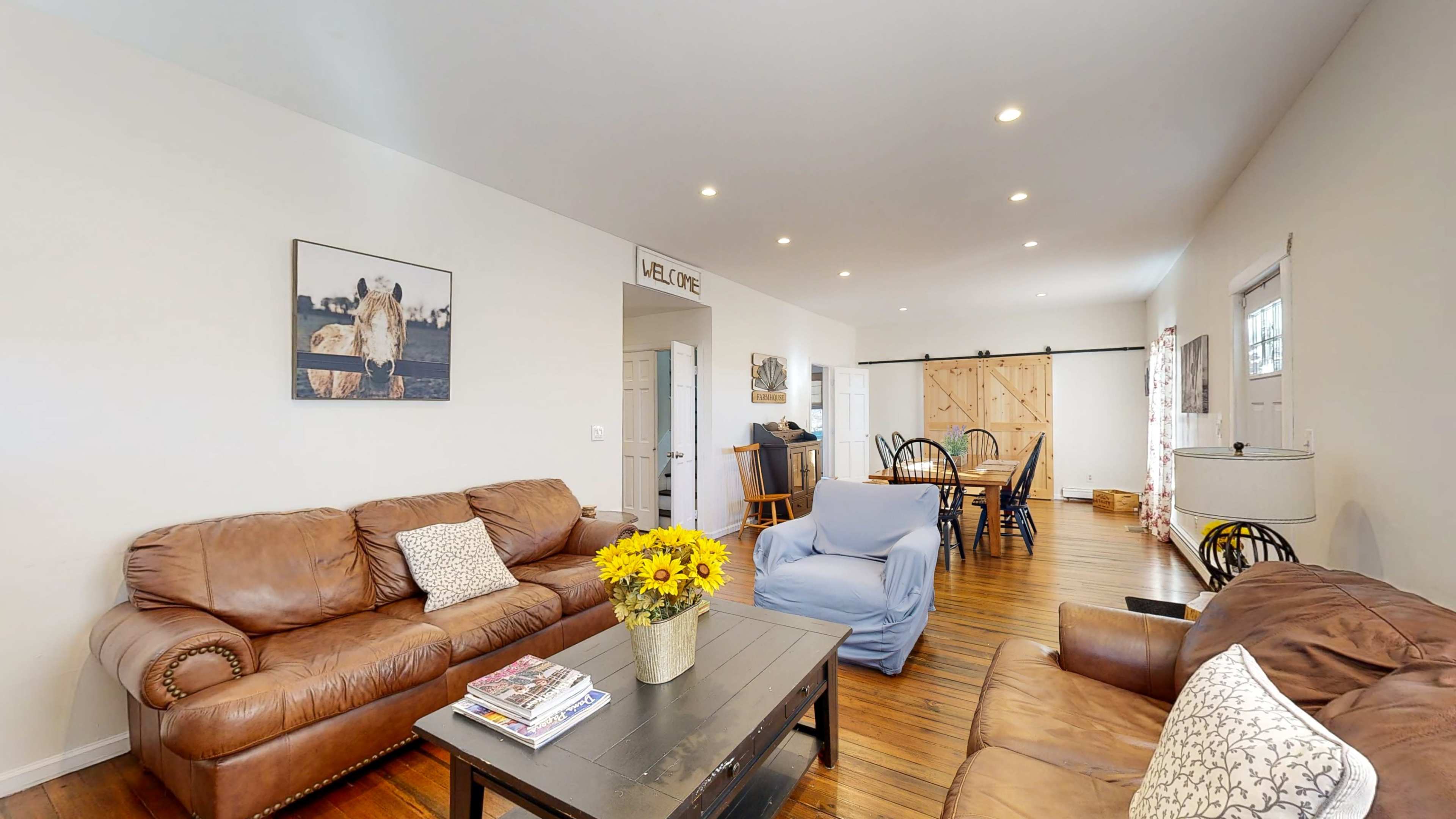 A cozy living area with leather sofas, a coffee table, and a dining space in the background featuring wooden floors and a large sliding door.