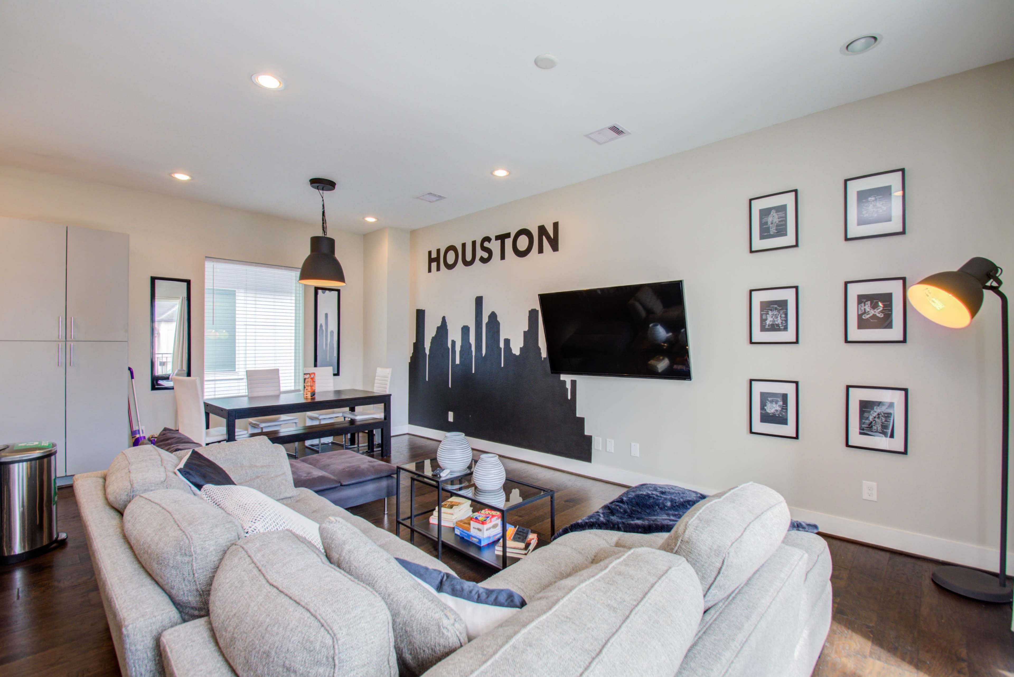 The living room features a gray sectional sofa, a coffee table, a television mounted on the wall, and a skyline mural of Houston above framed photographs.
