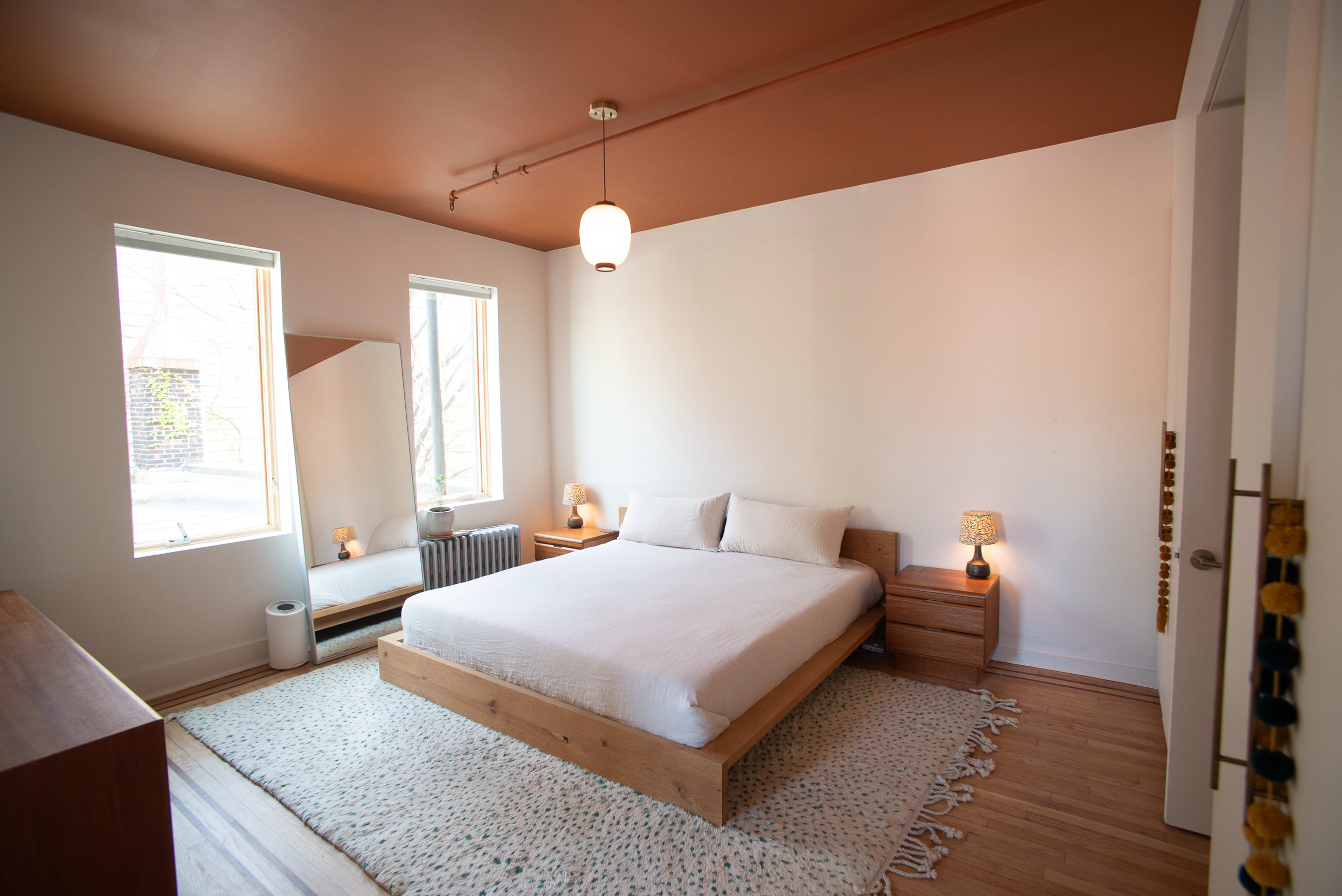 The image shows a minimalist bedroom featuring a wooden bed with white bedding, two bedside tables with lamps, and large windows allowing natural light to enter.